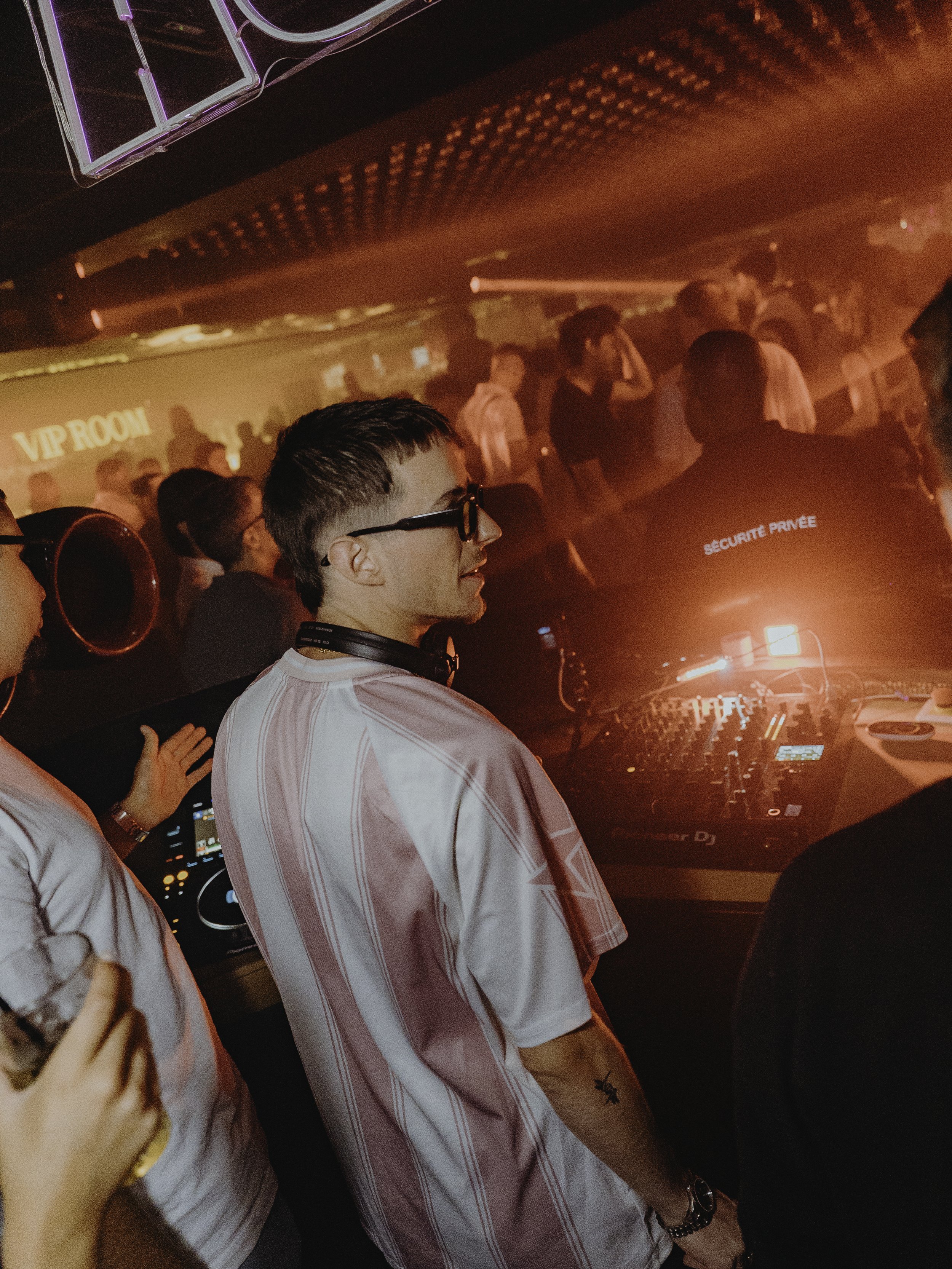 Jeune DJ avec lunettes et tatouage, à un concert ou une fête, entouré de personnes, ambiance rouge et sombre avec éclairage de club.