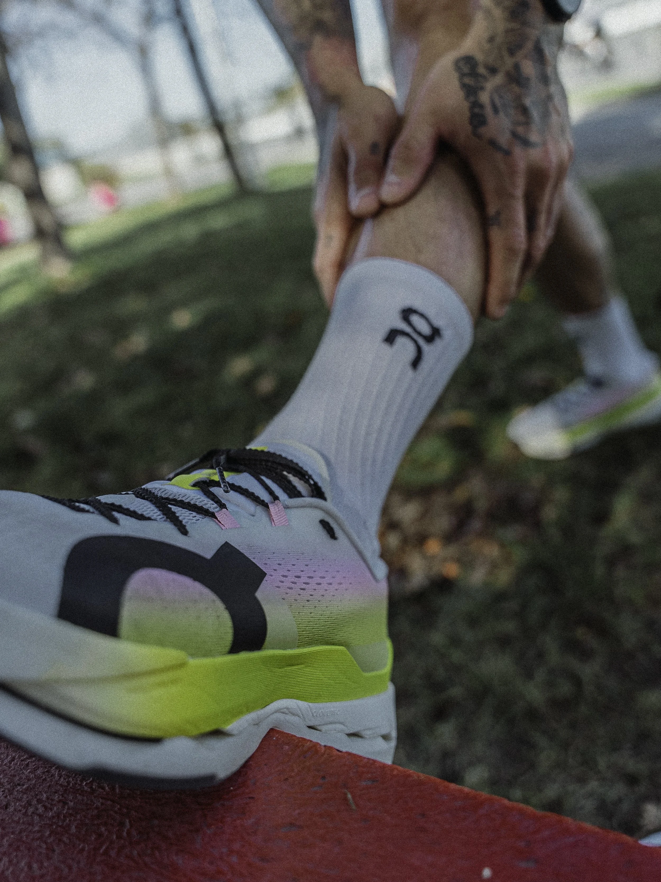 Une personne porte des baskets blanches avec des accents de couleur jaune, noir et rose, et des chaussettes blanches avec le logo 'OF'. La personne s'étire ou s'entretient avec une autre personne, dont la main est visible en train de saisir doucement