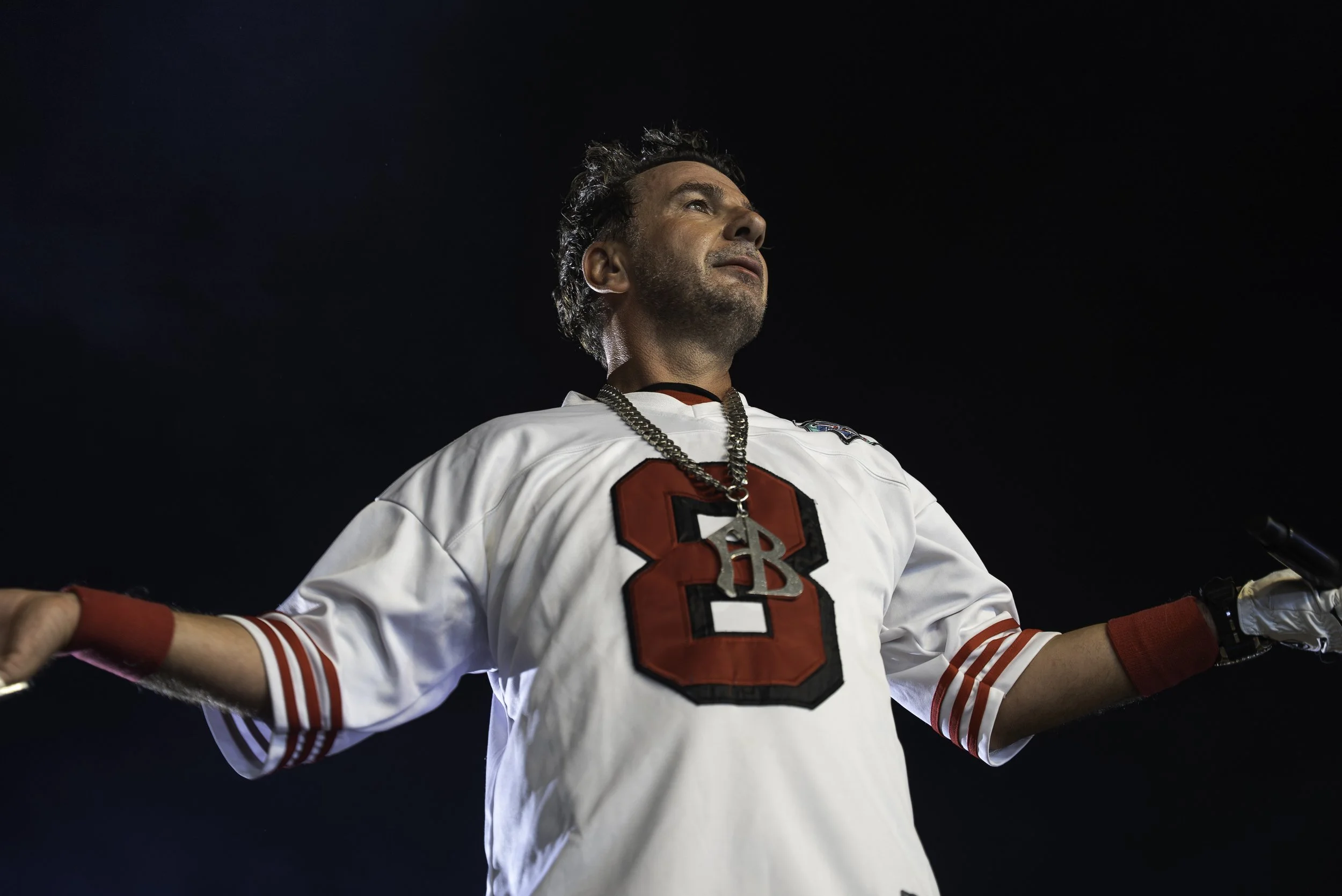 Un homme portant un maillot de football blanc avec un grand chiffre 3 rouge et noir sur la poitrine, un collier avec un médaillon TB, et des brassards rouges, se tient avec les bras ouverts, face à un ciel sombre.