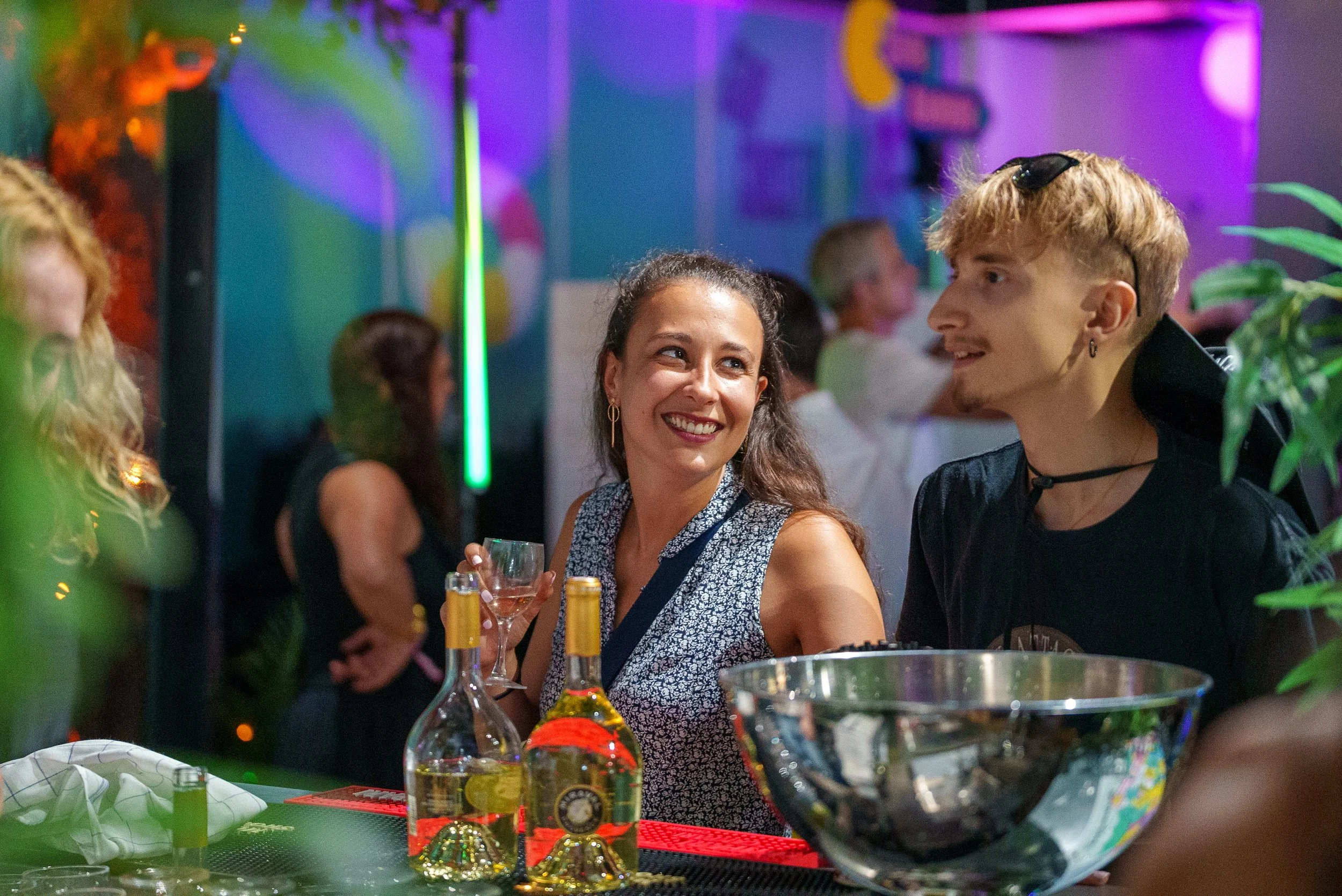 Deux personnes discutent et sourient dans un bar ou une boîte de nuit avec une ambiance colorée et lumineuse, des bouteilles d'alcool sur le comptoir.