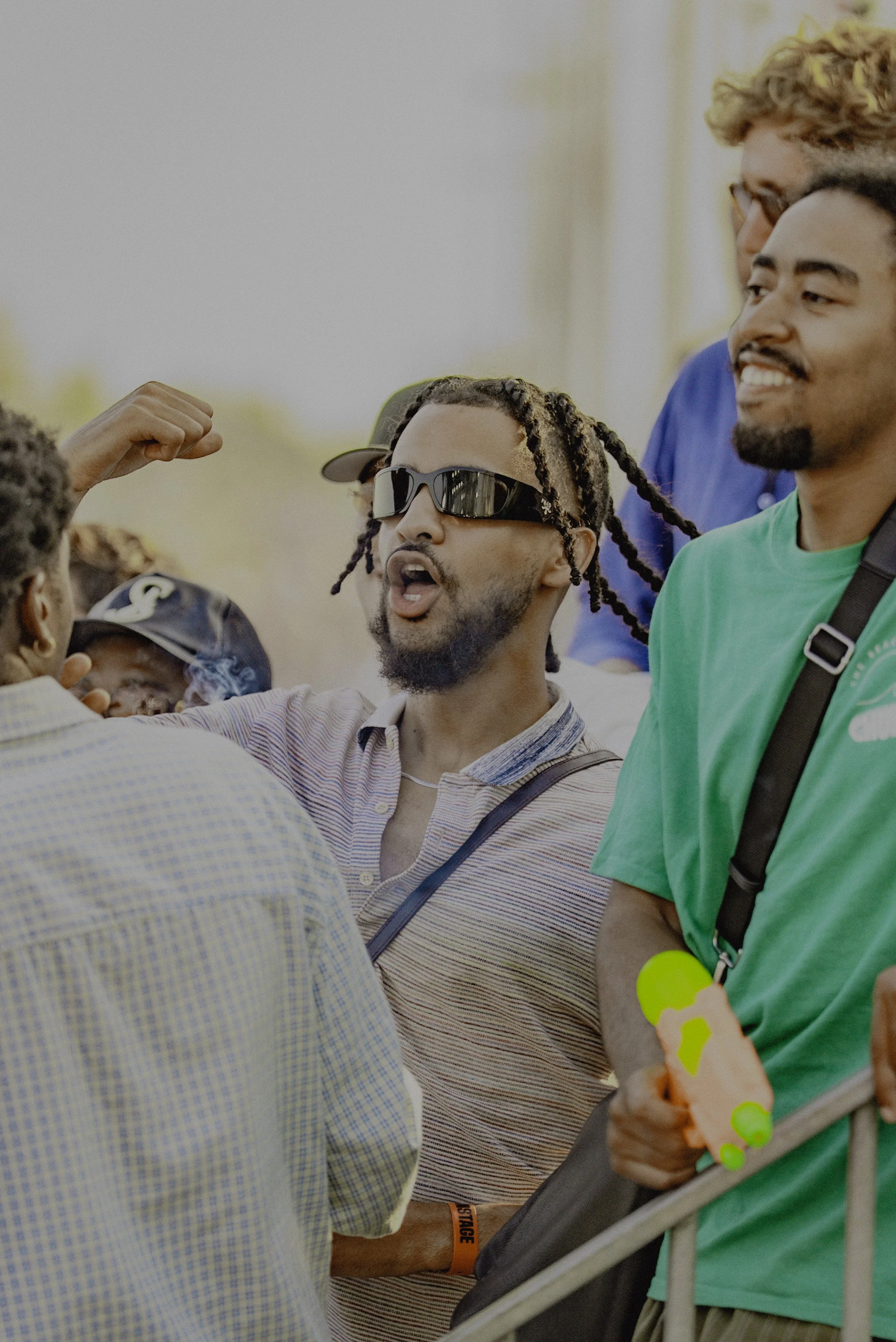 Un groupe de jeunes hommes discutent à l'extérieur, l'un d'eux portant des lunettes de soleil foncées et une chemise à rayures, semble s'engager dans une conversation animée.