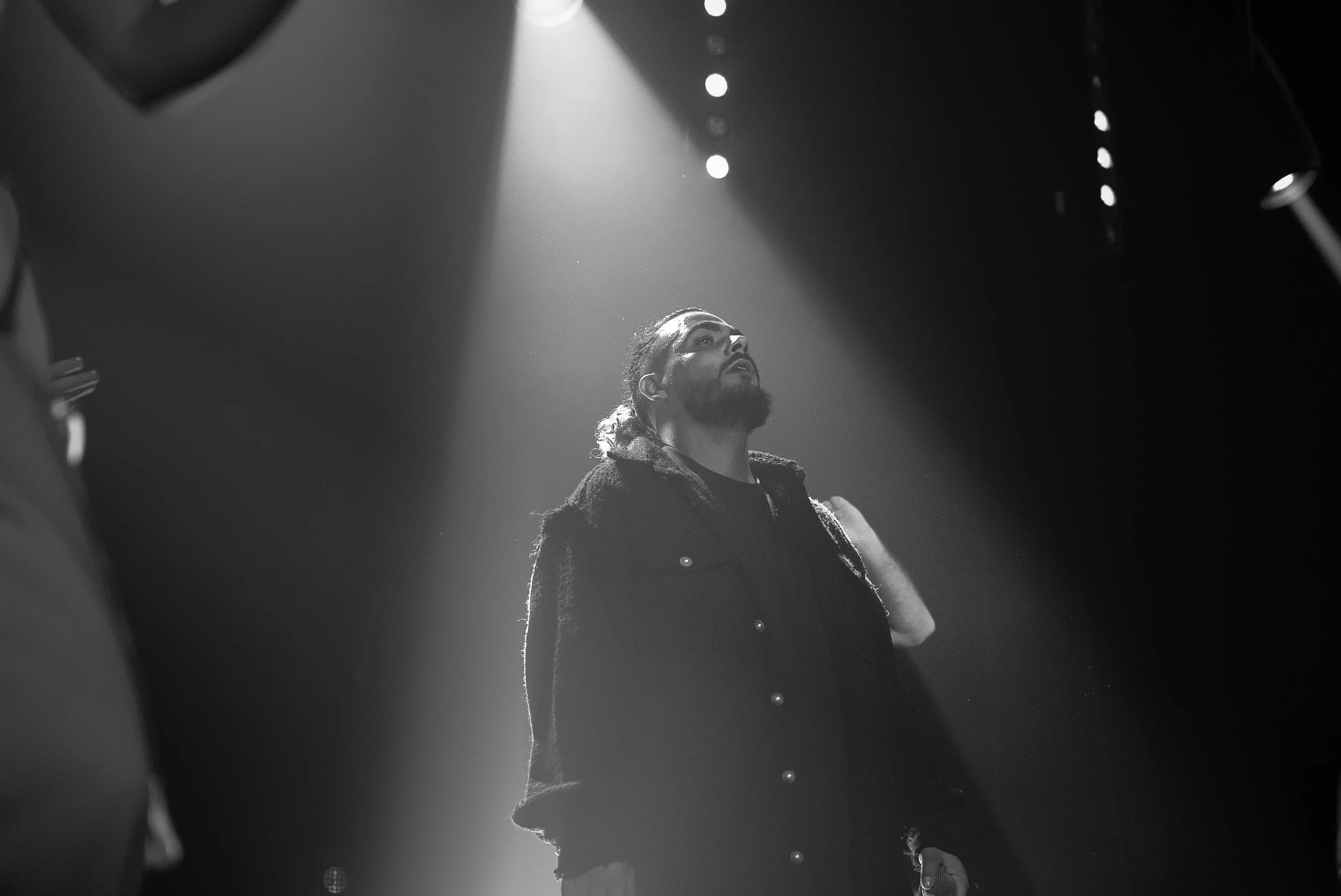 Un homme avec une barbe et des cheveux longs attachés, regardant vers le haut, sous un faisceau de lumière en scène ou en studio, en noir et blanc.