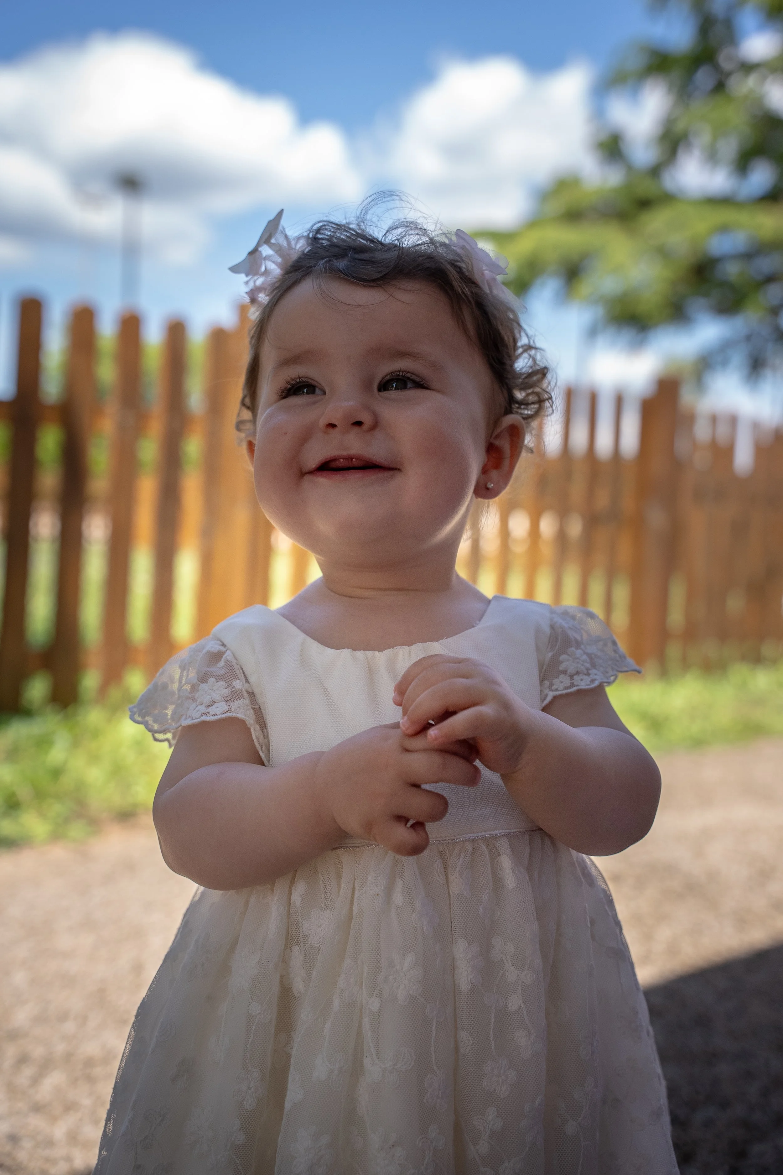 Jeune enfant souriante dehors, portant une robe blanche avec des détails en dentelle, derrière une clôture en bois, sous un ciel bleu avec des nuages.
