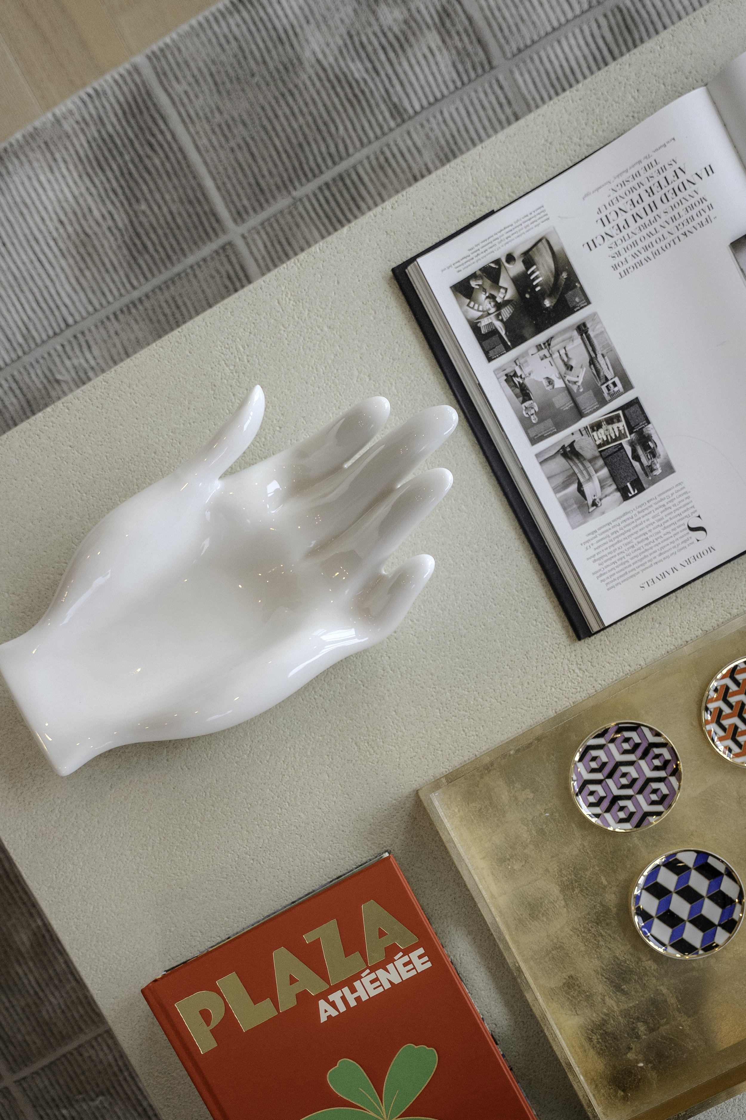 Une main en porcelaine blanche, un magazine ouvert avec des photos en noir et blanc, un livre rouge intitulé 'PLAZA ATHÉNÉE', et trois petits plats ronds avec des motifs géométriques colorés, posés sur une surface en bois et un tapis.