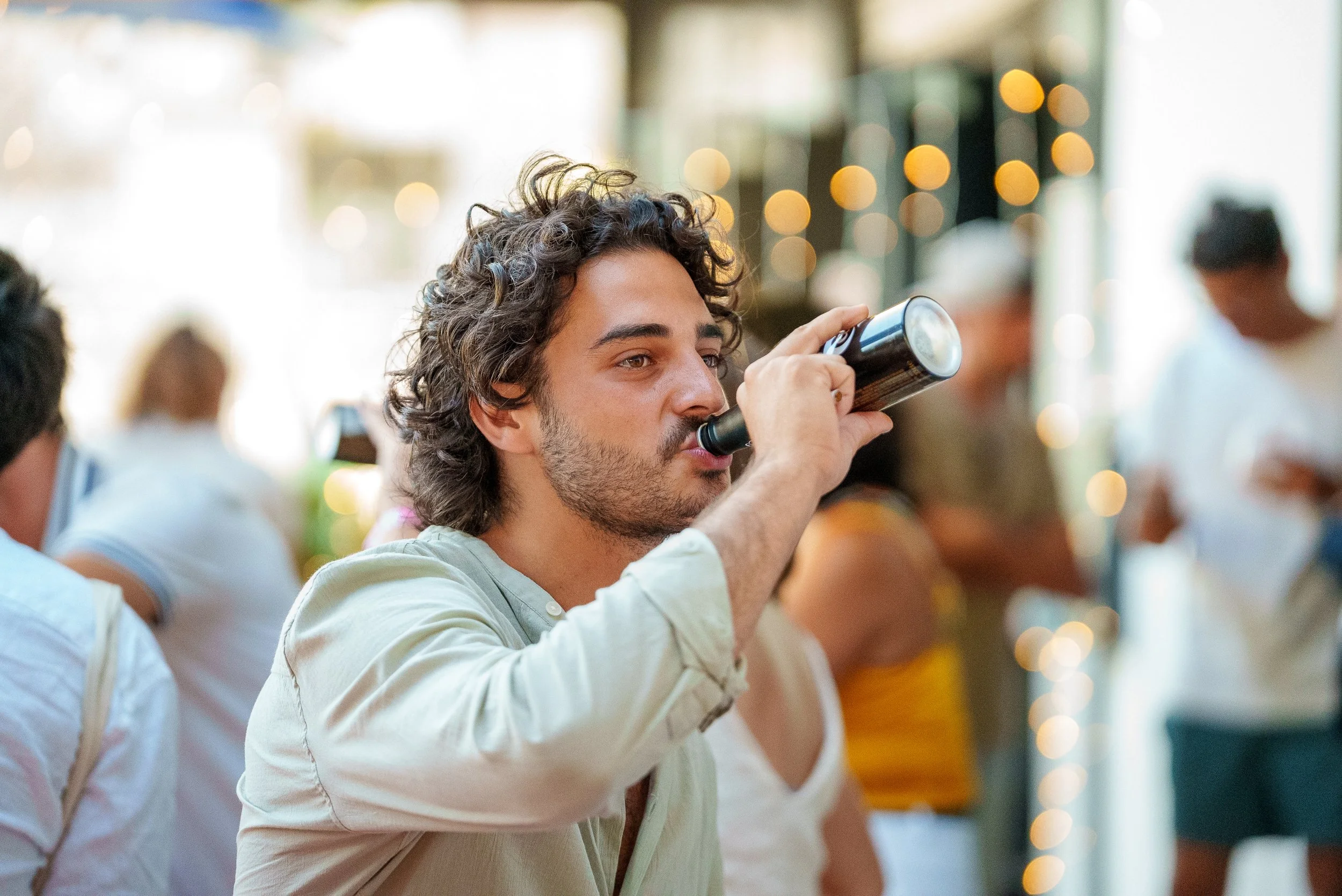 Un homme avec des cheveux bouclés et une barbe, en train de boire une bouteille de bière dans un endroit animé et lumineux.