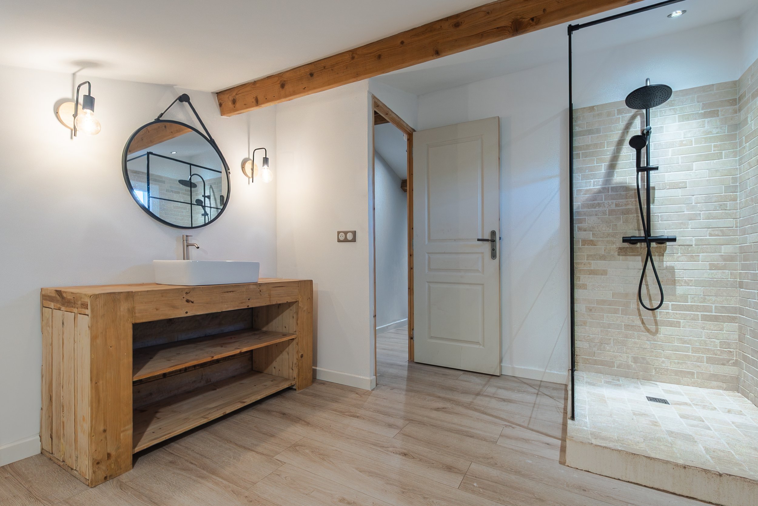 Salle de bain moderne avec lavabo en porcelaine sur meuble en bois, miroir rond, éclairage mural, espace douche avec paroi en verre et robinet noir.