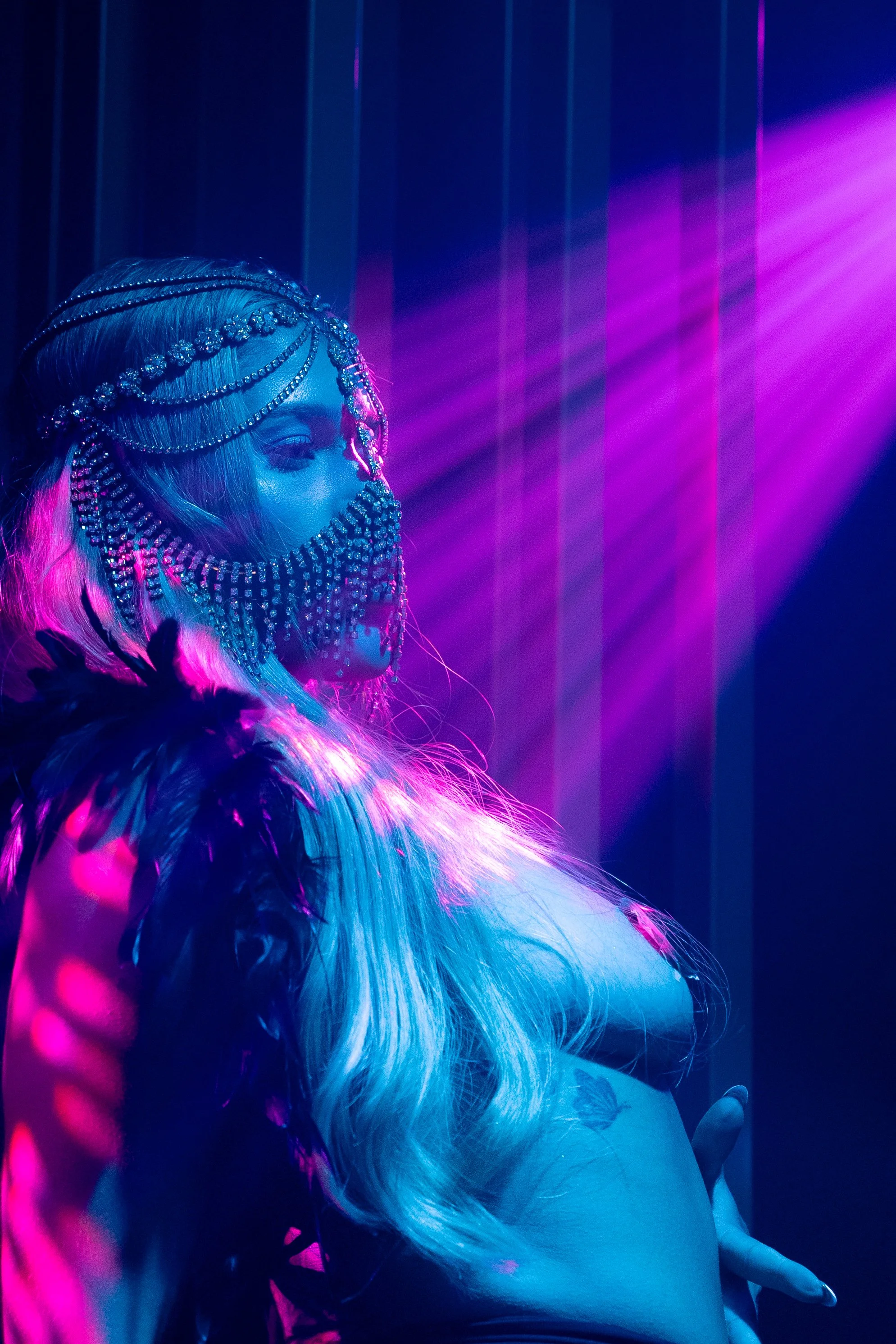 Une femme portant un bijou de tête étincelant et une robe de plumes, sous un éclairage violet et rose, dans une ambiance artistique.