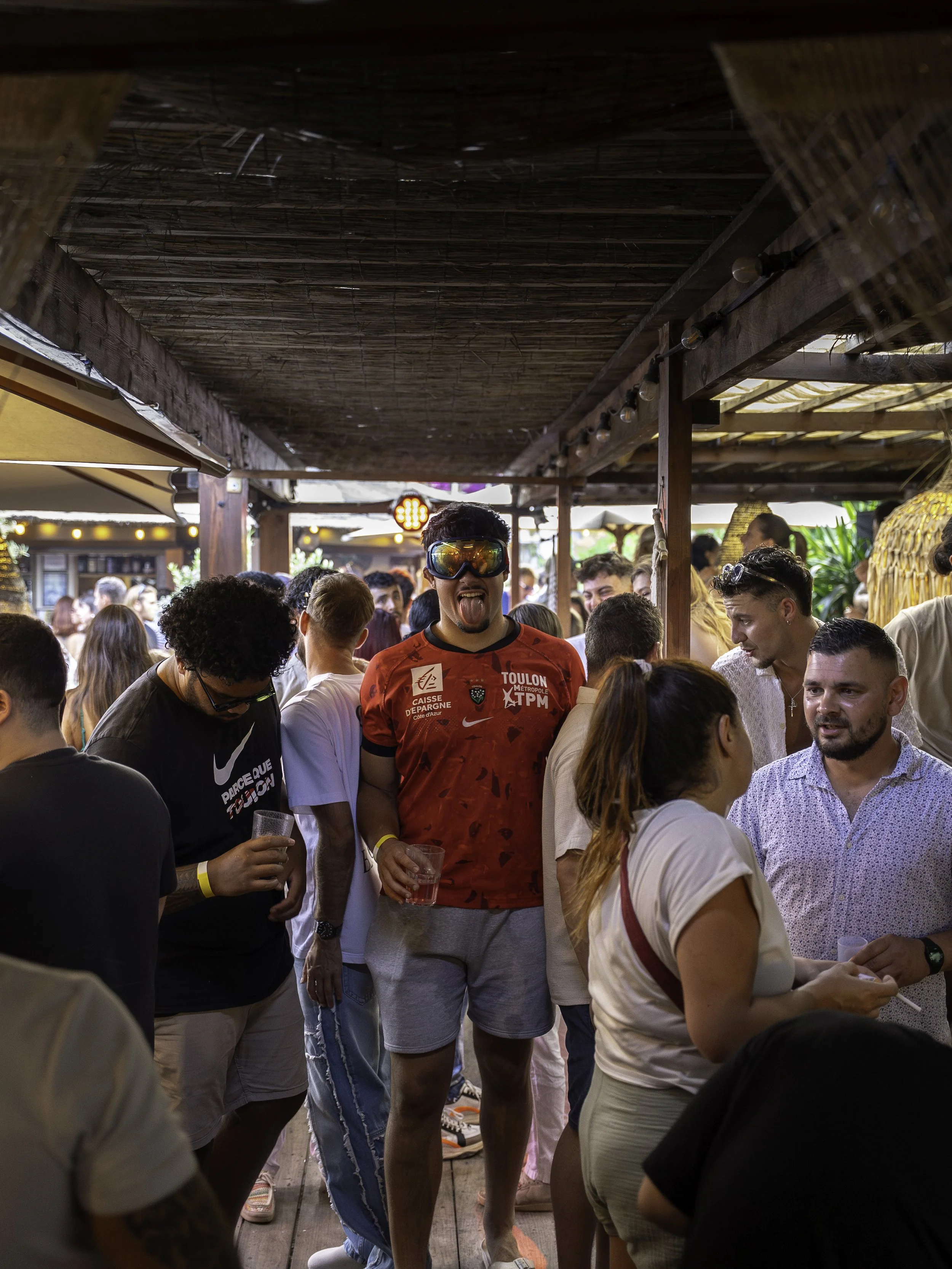 Groupe de personnes dans une ambiance festive à l'extérieur, avec un homme en lunettes de ski et maillot de sport en train de faire une grimace.