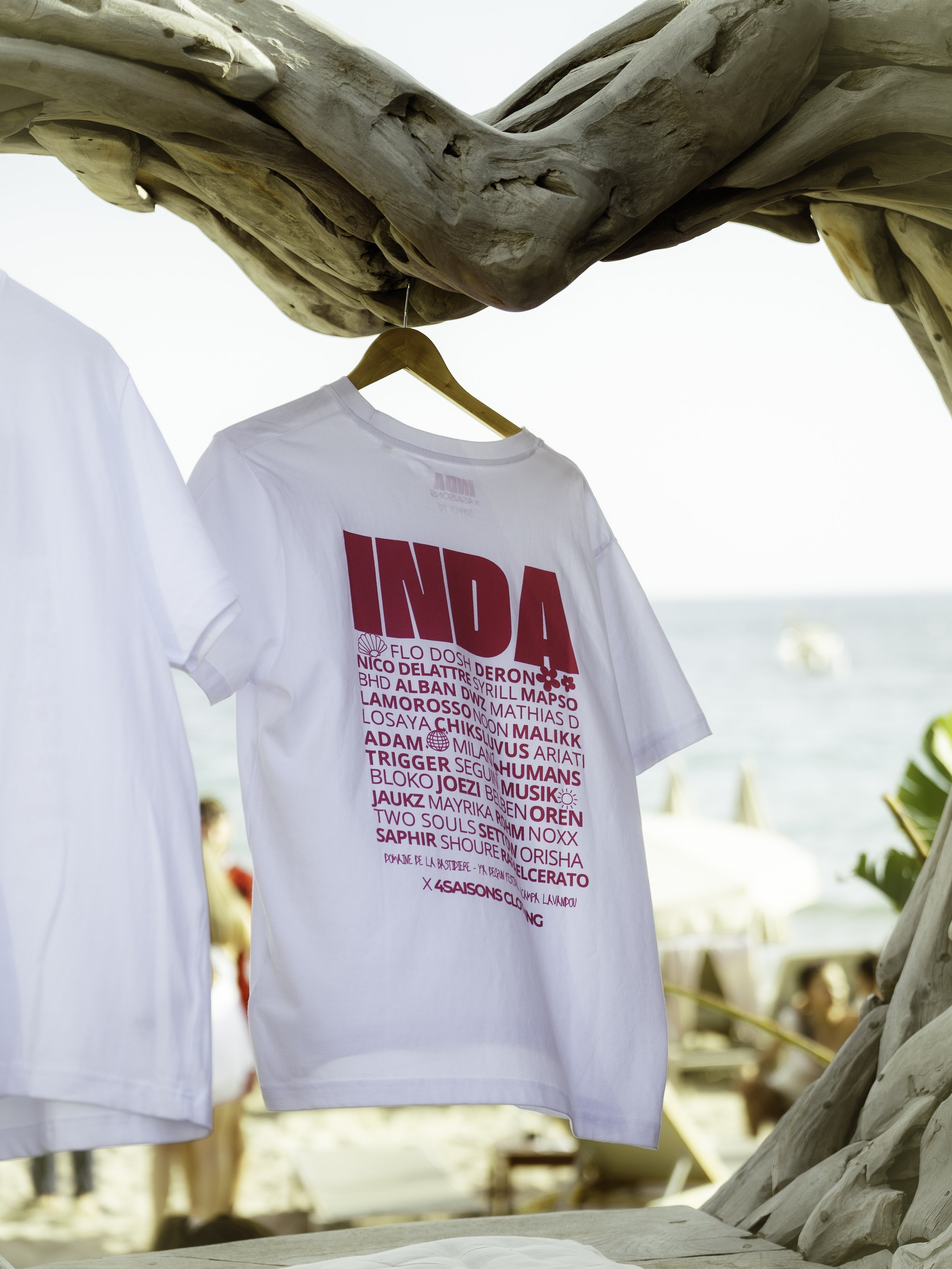 Une t-shirt blanc avec pratique en rose et en violet, accroché à un cintre en bois, suspendu à une structure en bois sculpté, sur une plage avec des personnes et la mer en arrière-plan.