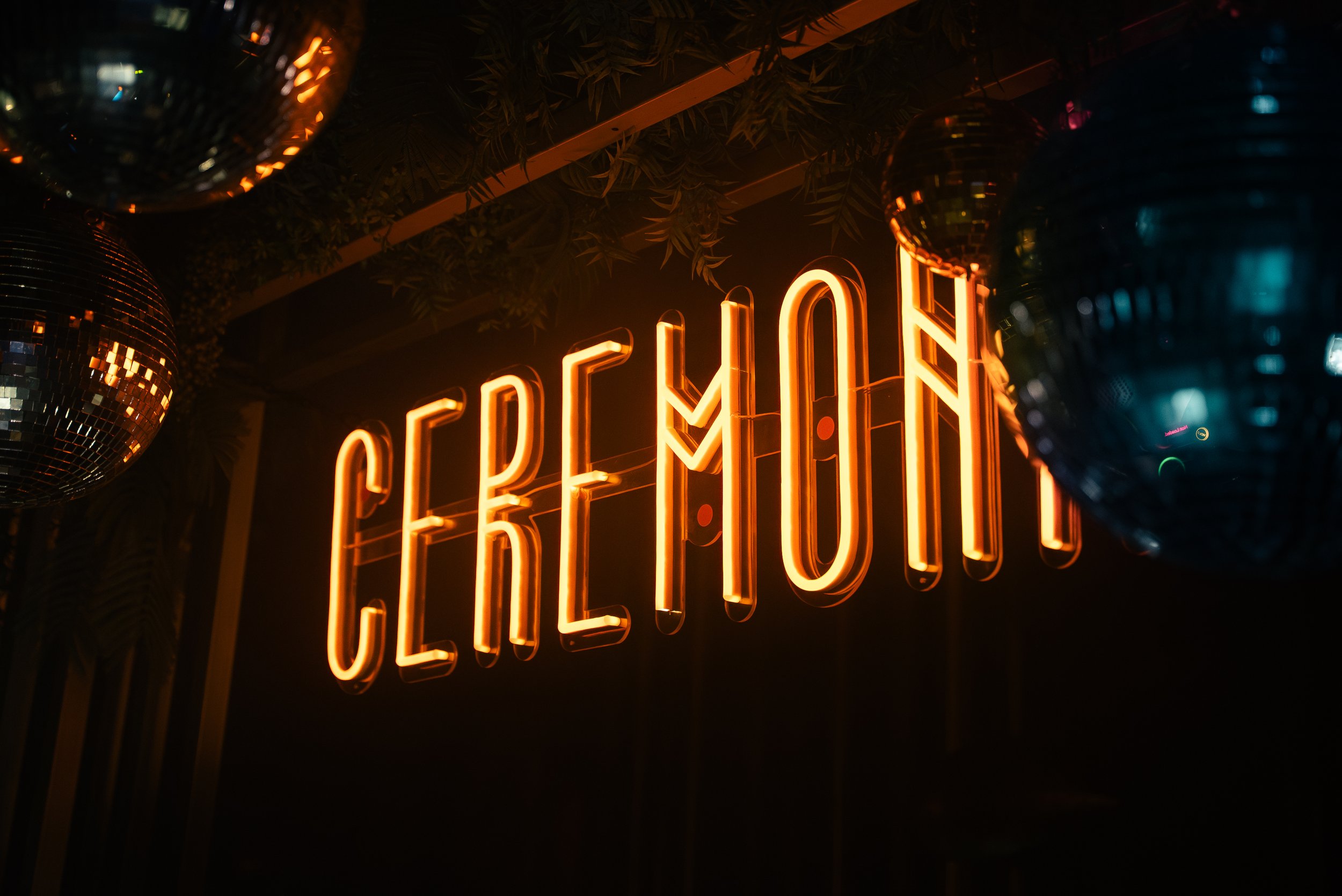 Un panneau lumineux orange avec le mot 'CÉREMONIE' écrit en lettres majuscules. Autour, il y a des ballons disco reflétant la lumière, dans un décor sombre et festif.