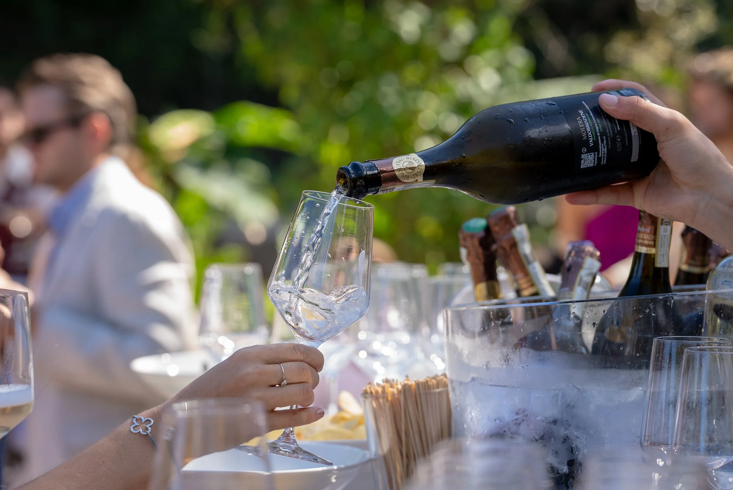 Une personne verse du vin dans un verre à l'extérieur, avec plusieurs bouteilles de champagne dans un seau à glace. Des personnes en arrière-plan portent des masques. L'ambiance semble être une dégustation ou un événement social.
