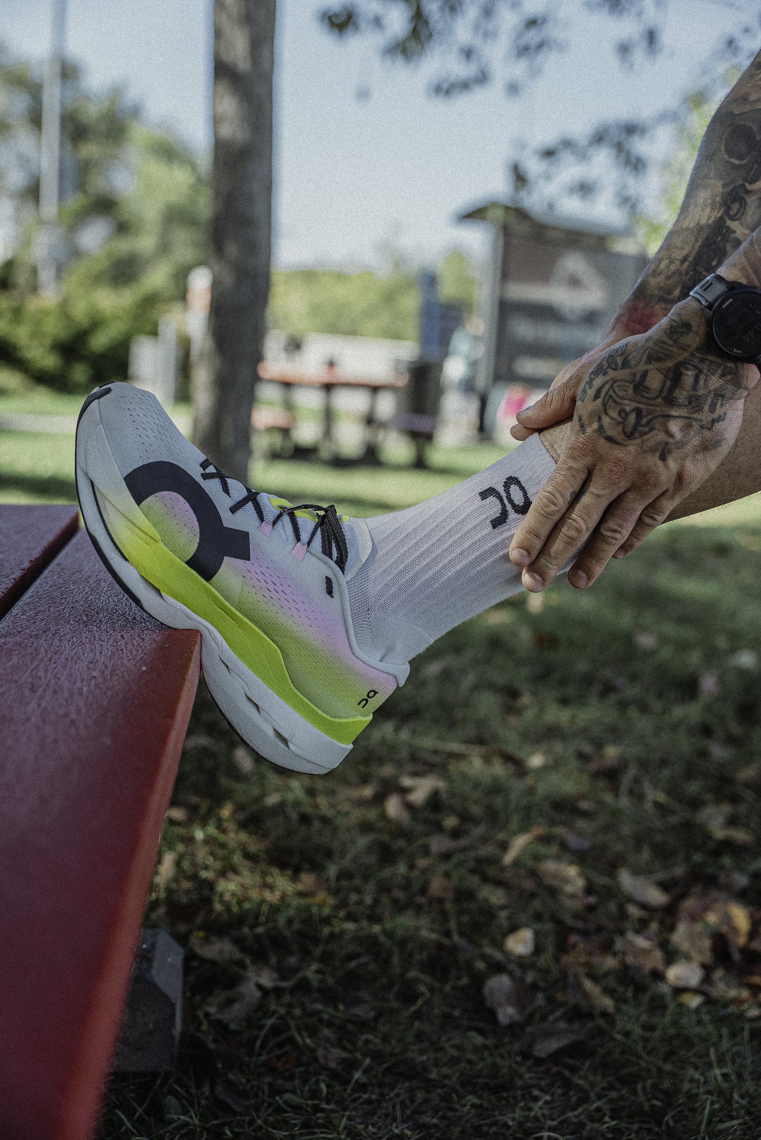 Une personne avec des tatouages aide à étirer la jambe d'un athlète portant une chaussure de course blanche avec des détails en jaune et rose, dans un parc en plein air.