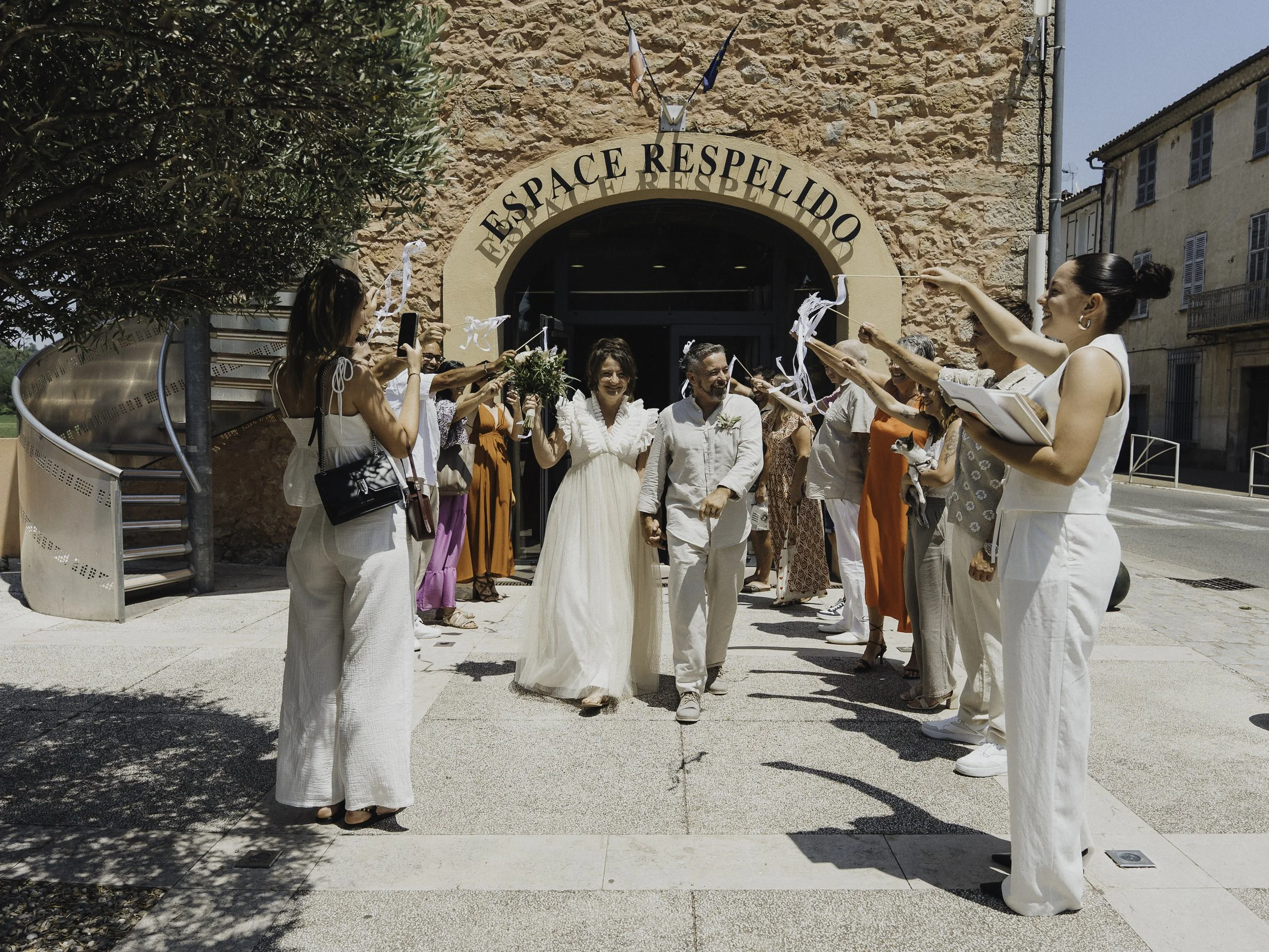 Groupe de personnes souriantes assais lors d'une célébration devant une entrée de bâtiment, certains tenant des confettis en l'air.