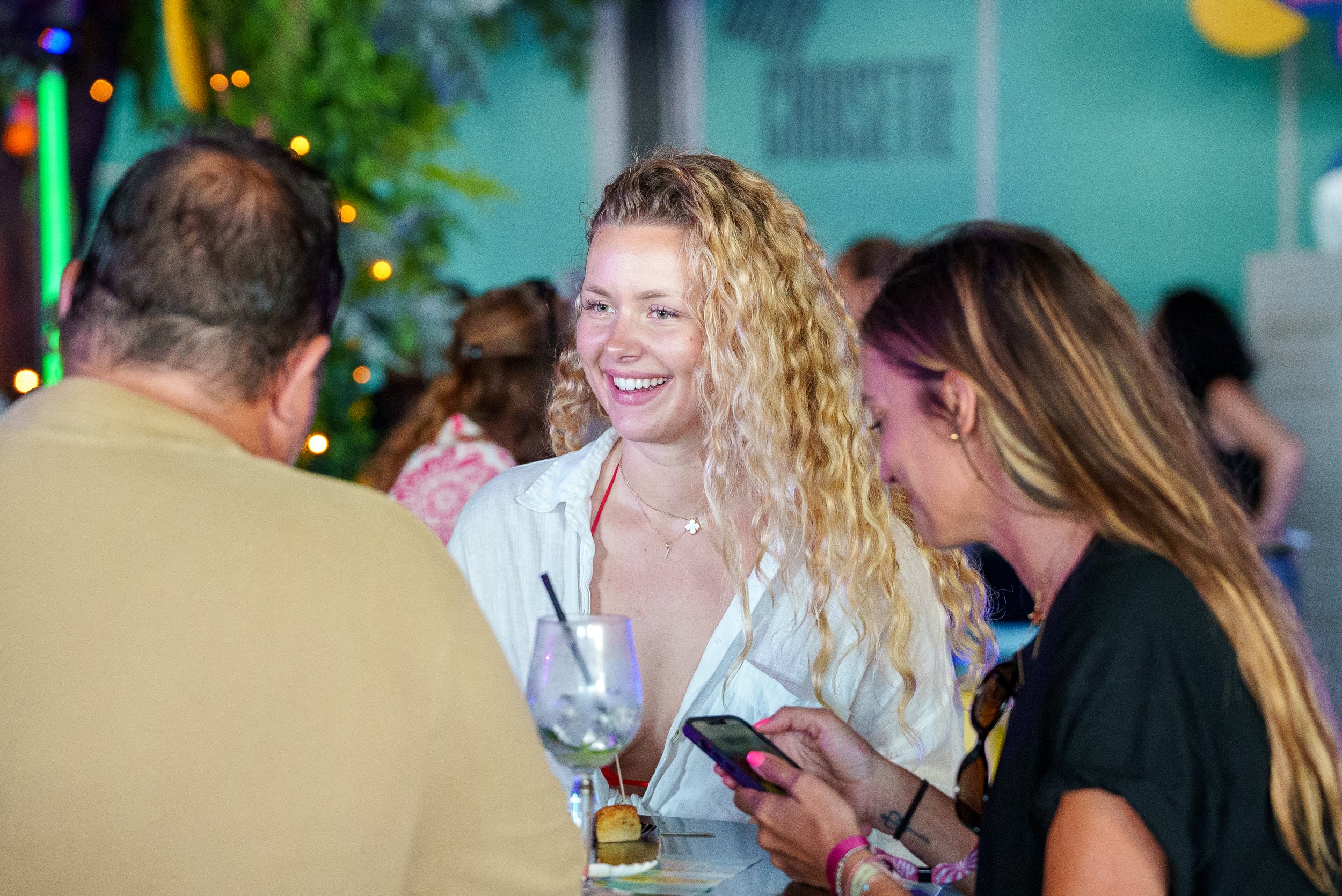 Trois personnes assises à une table de bar ou café, deux femmes et un homme, en train de converser, avec une boisson et une petite assiette, dans un décor animé avec des lumières colorées en arrière-plan.