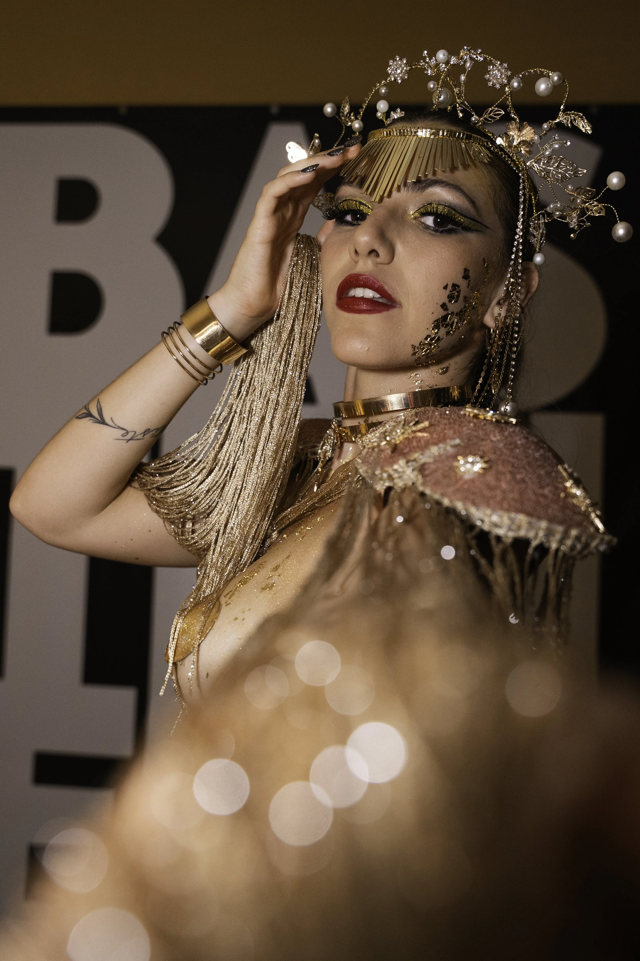 Une femme avec un maquillage sophistiqué, portant une tenue dorée et des accessoires ornés de bijoux, posant avec confiance, avec un fond noir et des lumières floues.