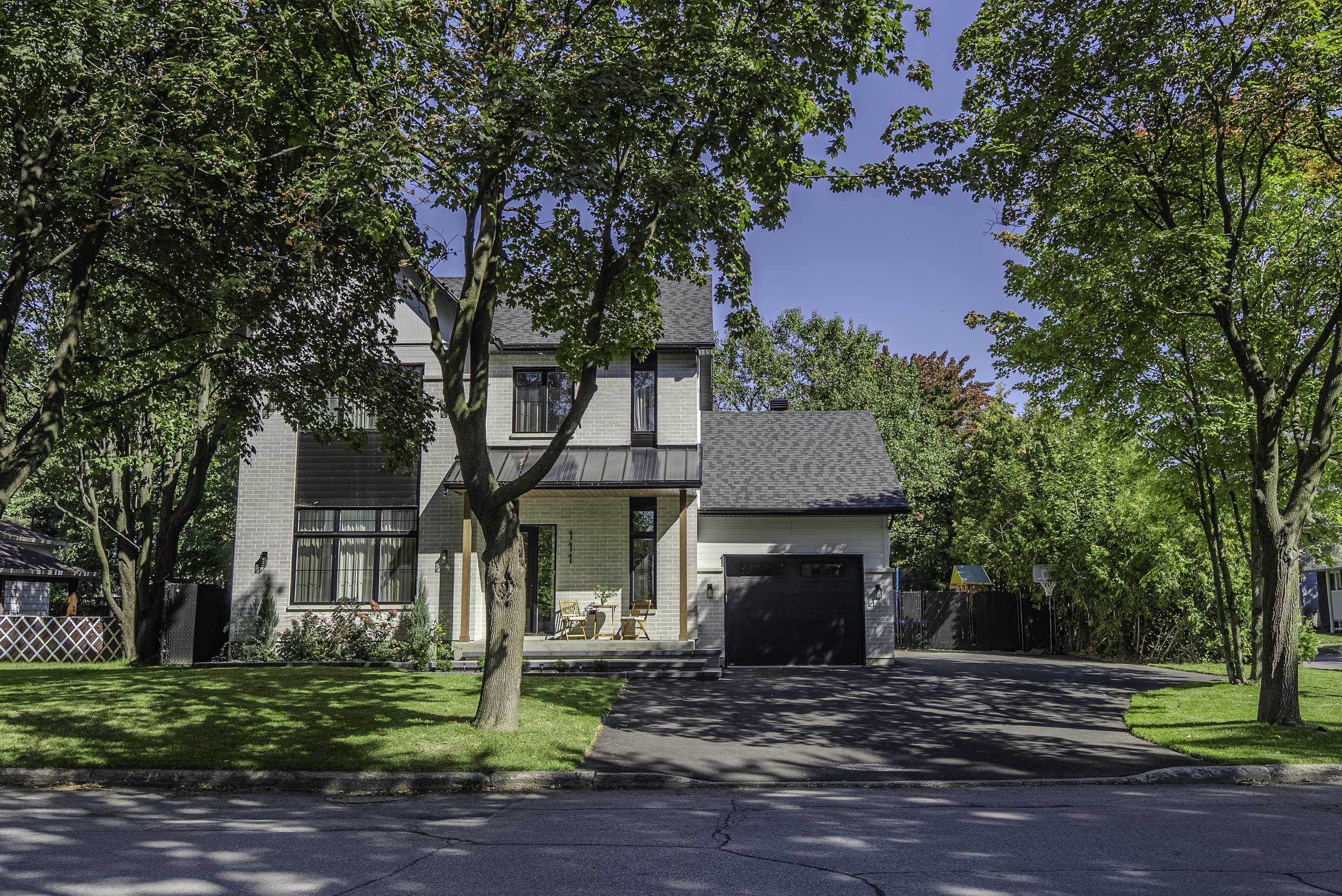 Maison moderne à deux étages avec garage, entourée d'arbres et d'une pelouse verte, sous un ciel bleu clair.
