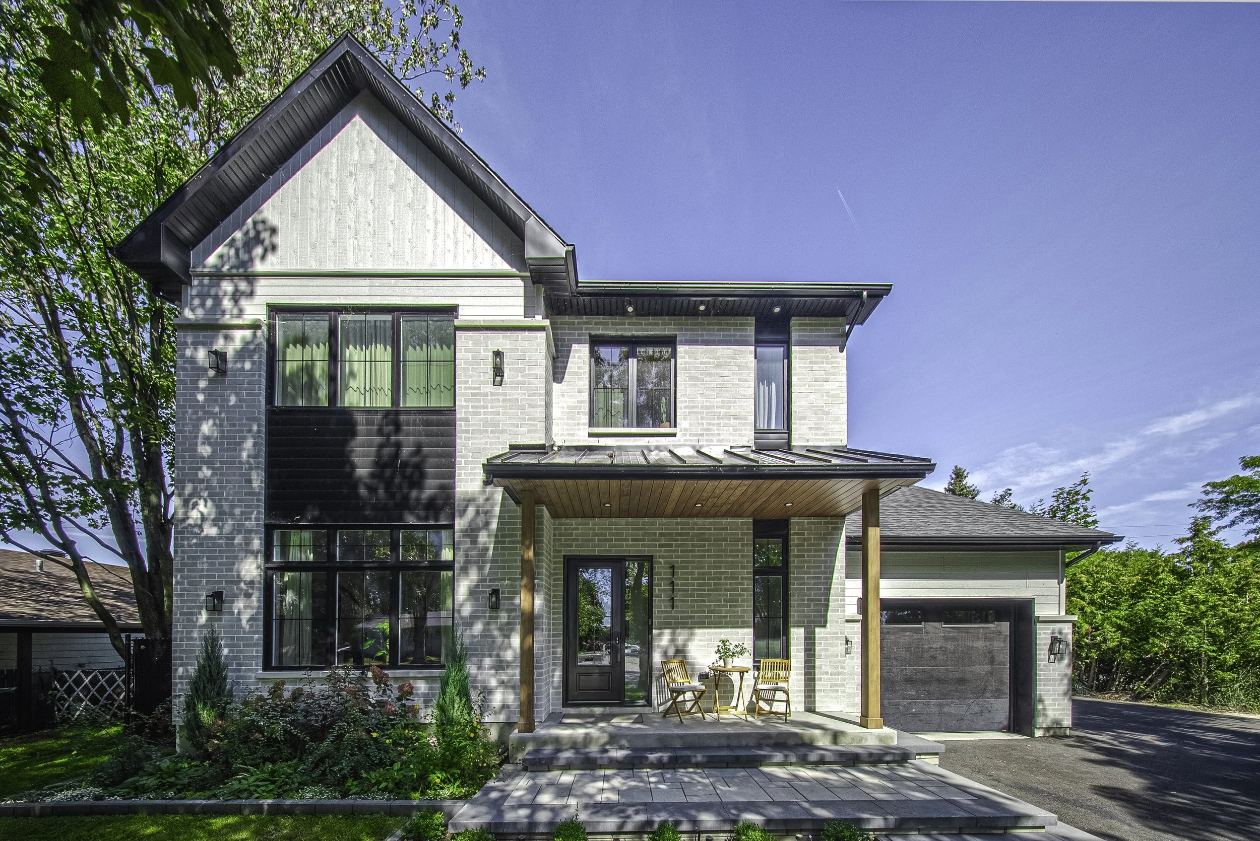 Maison moderne à deux étages avec cour pavée, jardin avec plantes, porte d'entrée en verre, garage, et façade en briques de couleur claire, sous un ciel bleu avec arbres environnants.