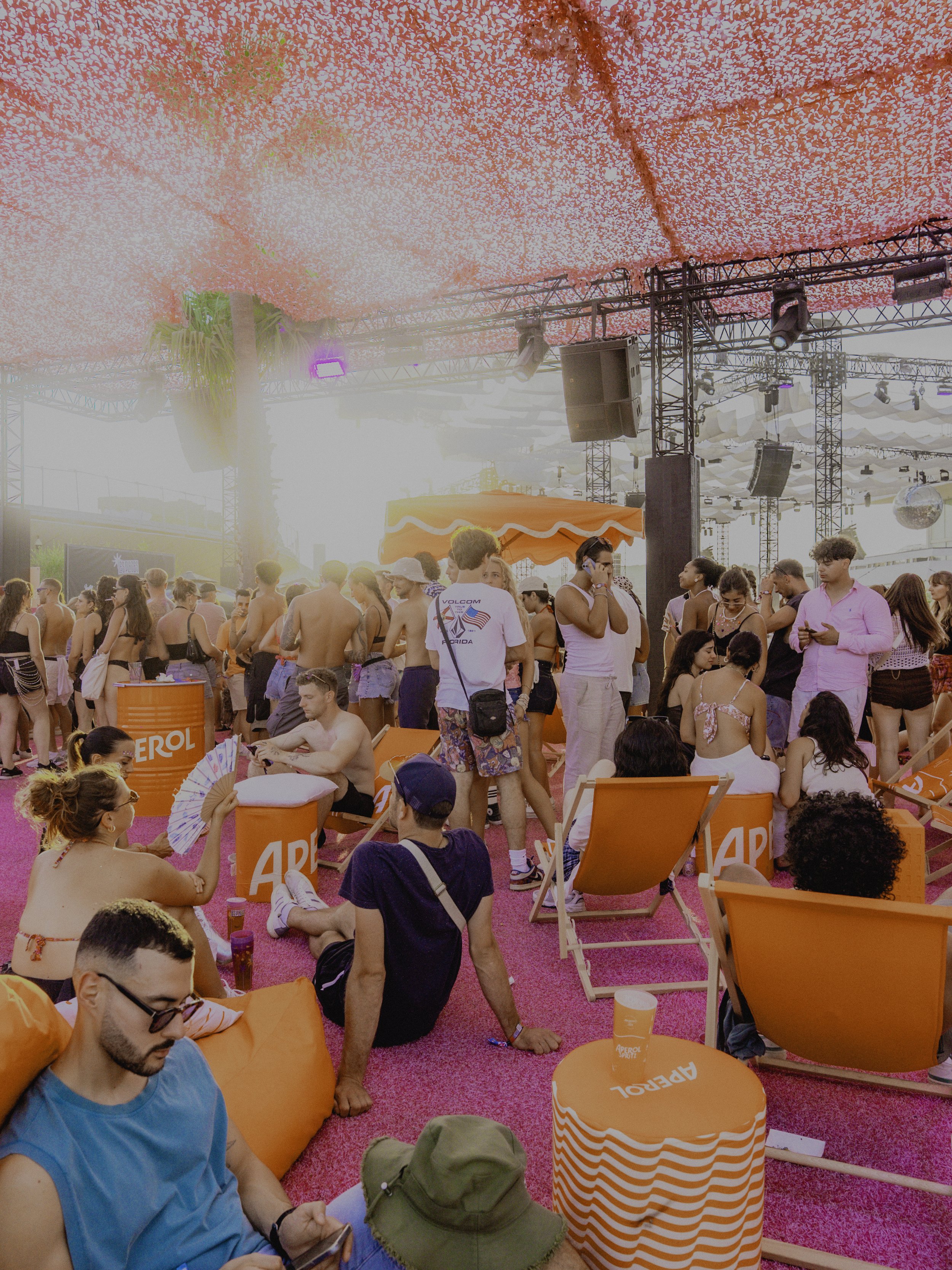 Groupes de personnes assises ou debout dans un espace de fête en plein air avec une décoration orange et rose, un toit de guirlandes roses et un éclairage. Certains personnes sont en maillot de bain, d'autres discutent ou utilisent leur téléphone.