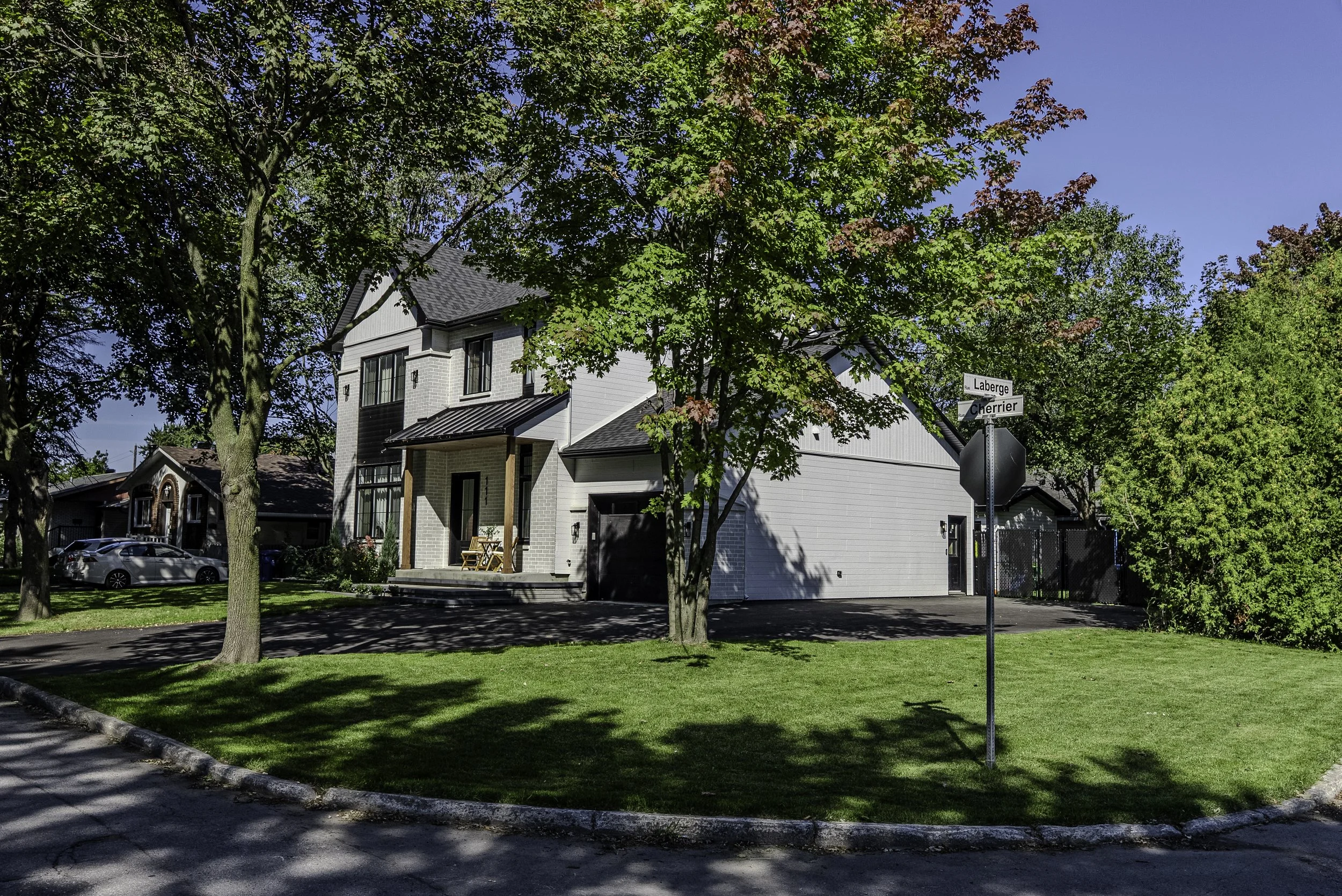 Maison moderne à deux étages avec garage noir, arbres feuillus, pelouse verte, ciel bleu en arrière-plan, panneaux de rue indiquant Laberge et Cherrier, et voiture dans la rue.