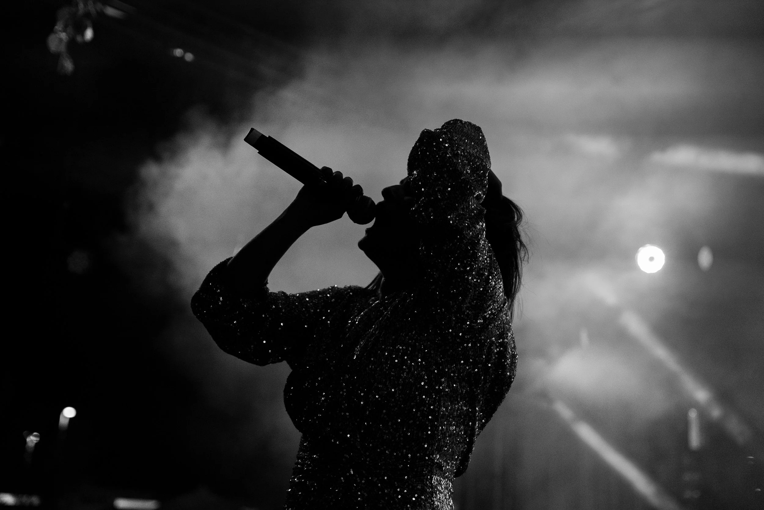 Silhouette d'une femme chantant avec un microphone lors d'un concert, portant un vêtement et un chapeau brillants, en noir et blanc.