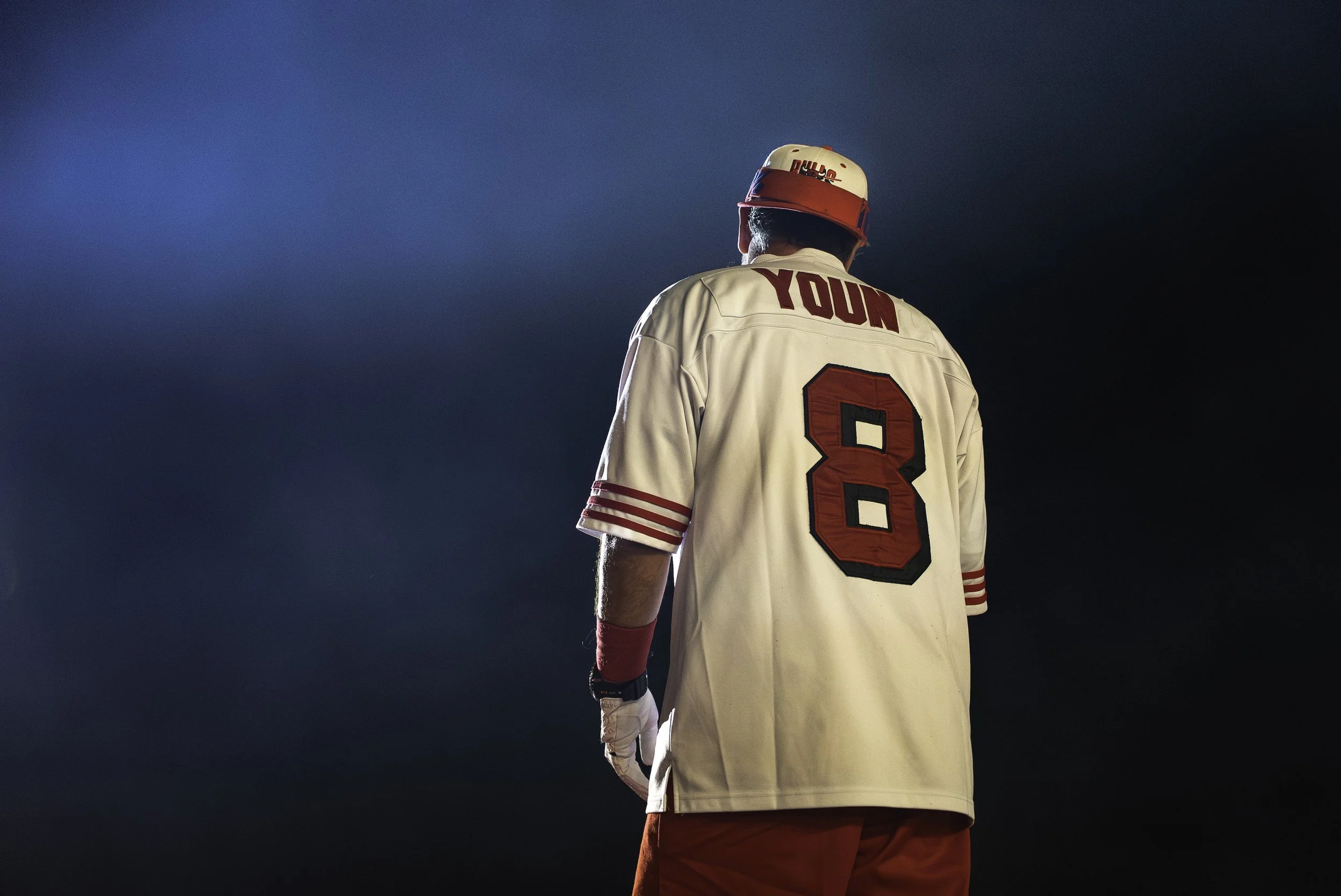 Un joueur de baseball vêtu d'un maillot blanc avec le nom 'YOUNG' et le numéro 8 à l'arrière, portant un casque blanc et rouge, vu de dos dans un éclairage dramatique