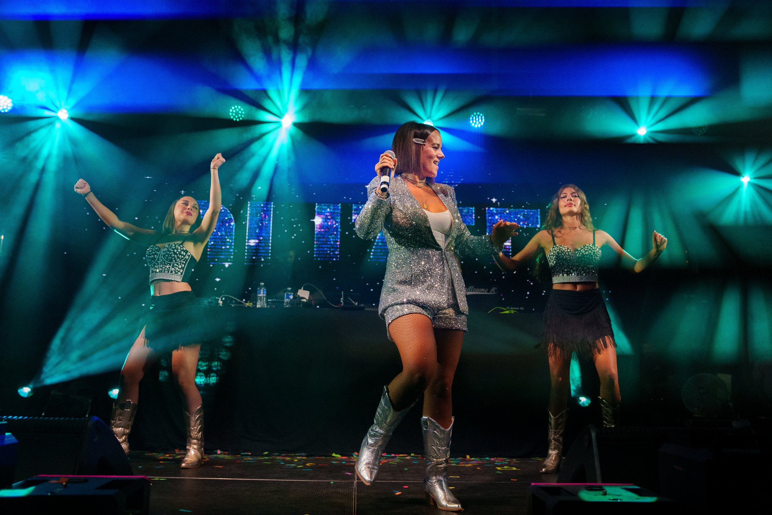 Une femme chante ou danse sur scène entourée de deux autres femmes, toutes en tenues brillantes, avec des lumières colorées et un fond numérique.