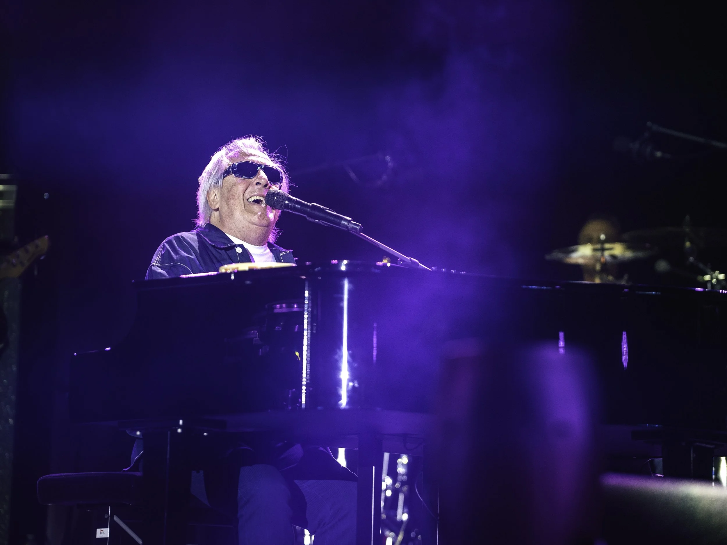 Un homme âgé avec des lunettes de soleil chante en jouant du piano lors d'une performance musicale sur scène, avec un fond sombre et des lumières bleues