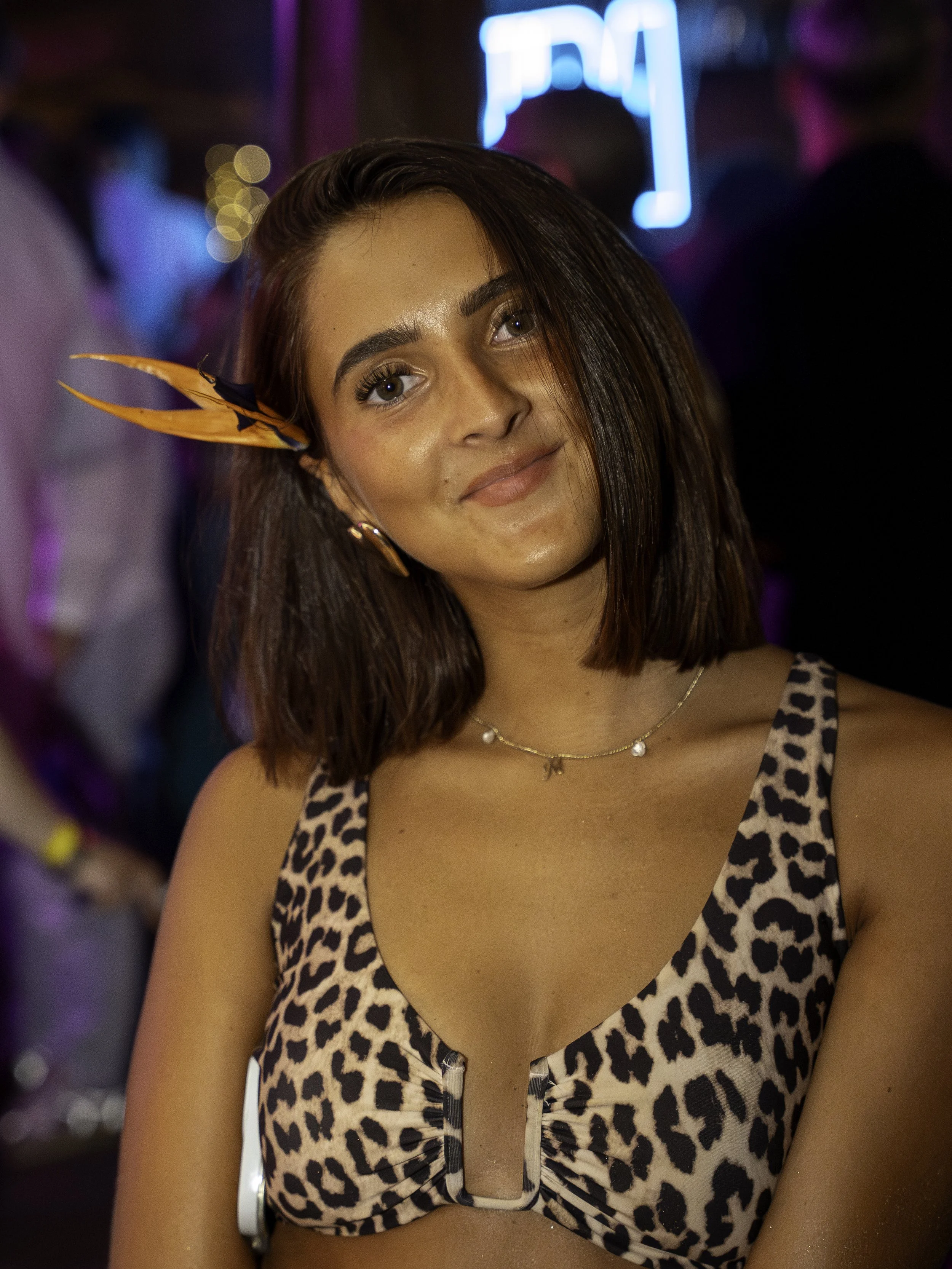 Jeune femme avec un bandeau orange et noir, portant un haut à motif léopard, souriant en soirée avec des lumières colorées en arrière-plan.