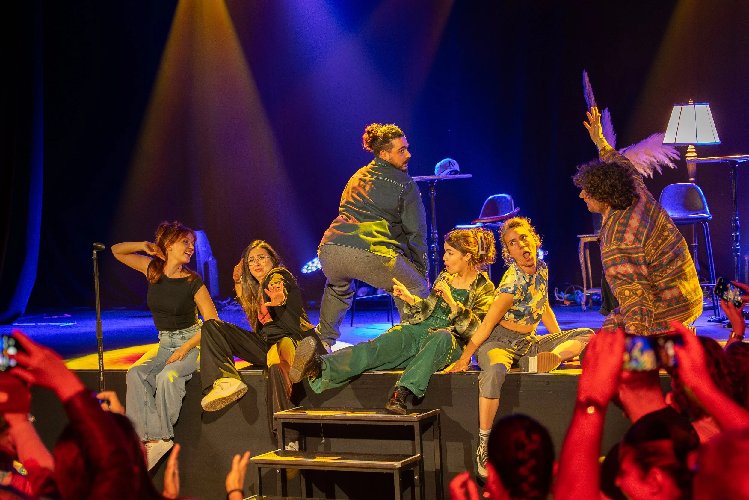 Groupe de personnes sur scène en train de jouer une pièce de théâtre, avec mise en scène colorée et spectateurs en avant-plan.