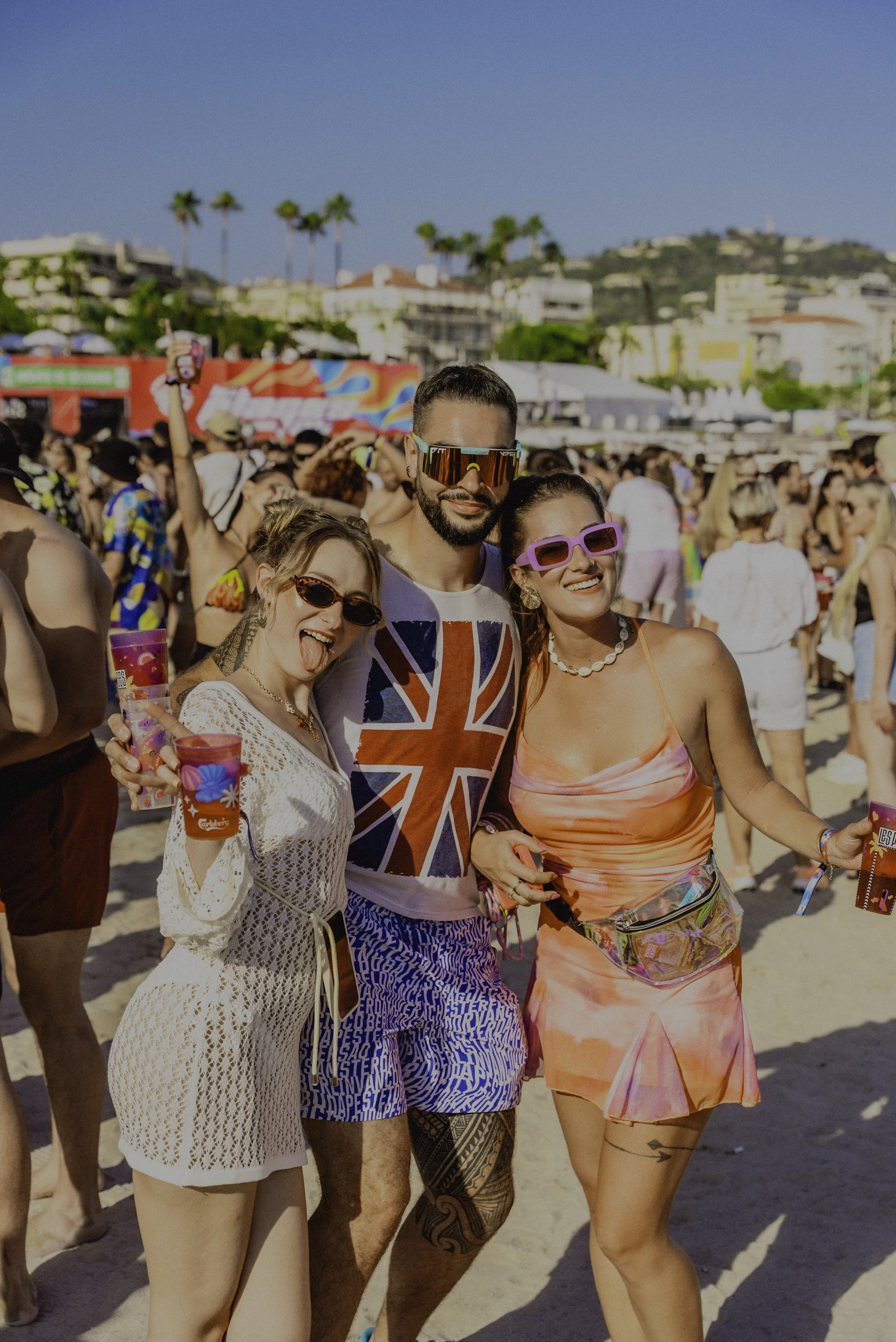 Groupe de trois personnes posant pour une photo lors d'un festival à la plage, avec beaucoup de gens en arrière-plan, tous souriants et habillés en vêtements d'été colorés.