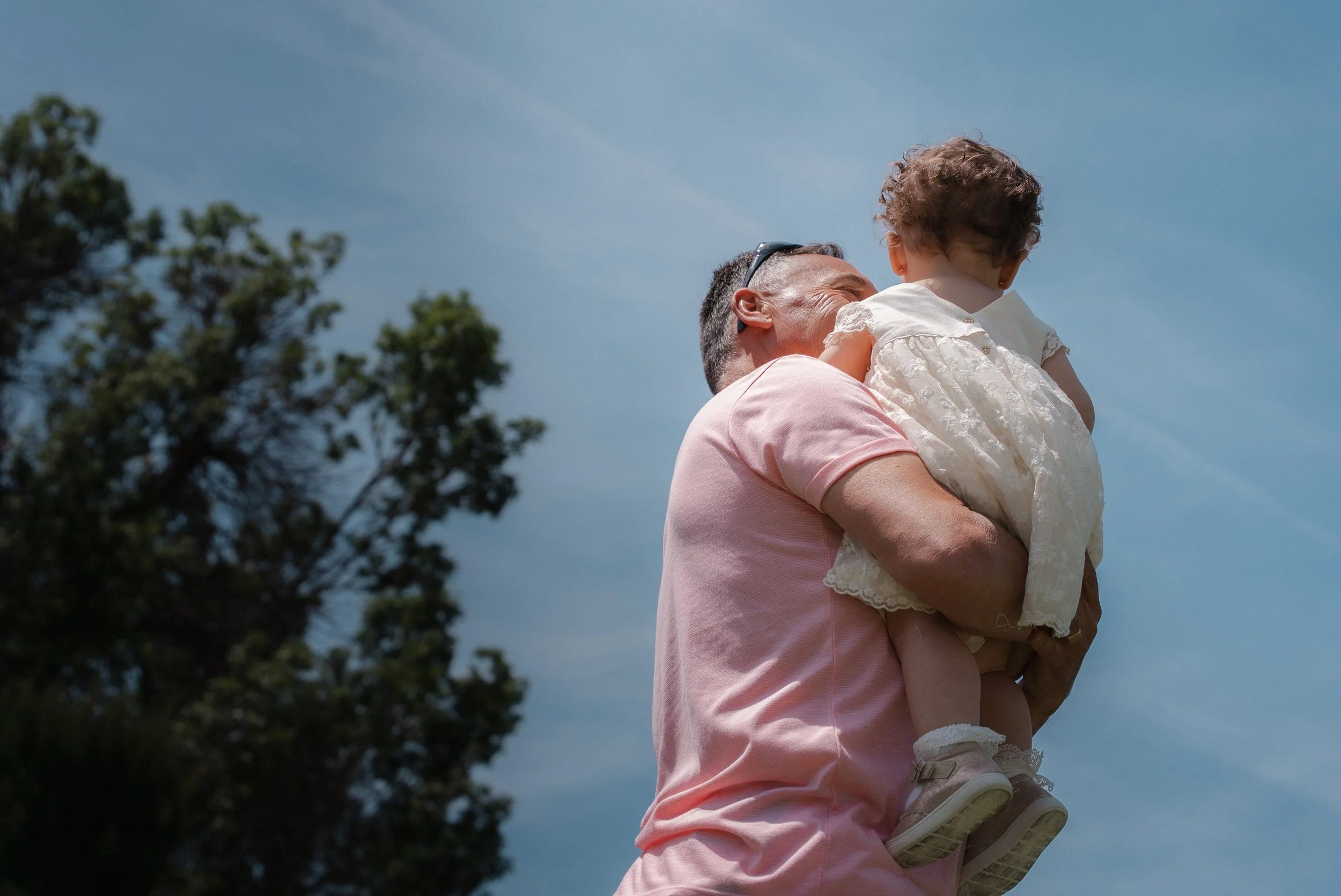 Un homme portant une chemise rose tient une jeune fille dans ses bras en plein air, le ciel bleu en arrière-plan.