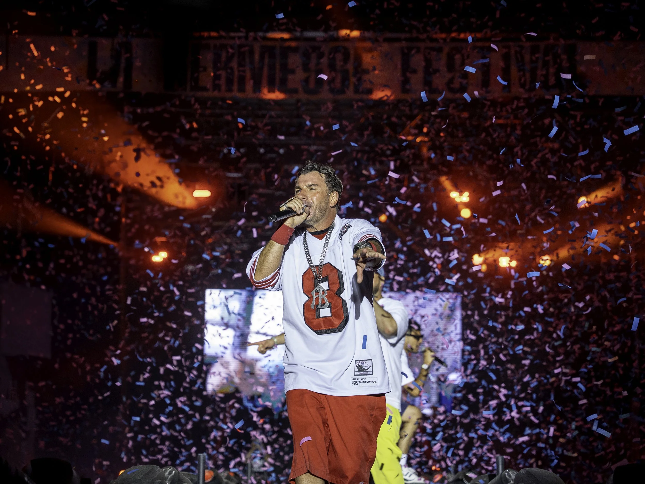 Un homme chante avec un microphone lors d'un concert, portant un maillot de football blanc avec un numéro 8 rouge, entouré de confettis colorés et de lumières rouges.