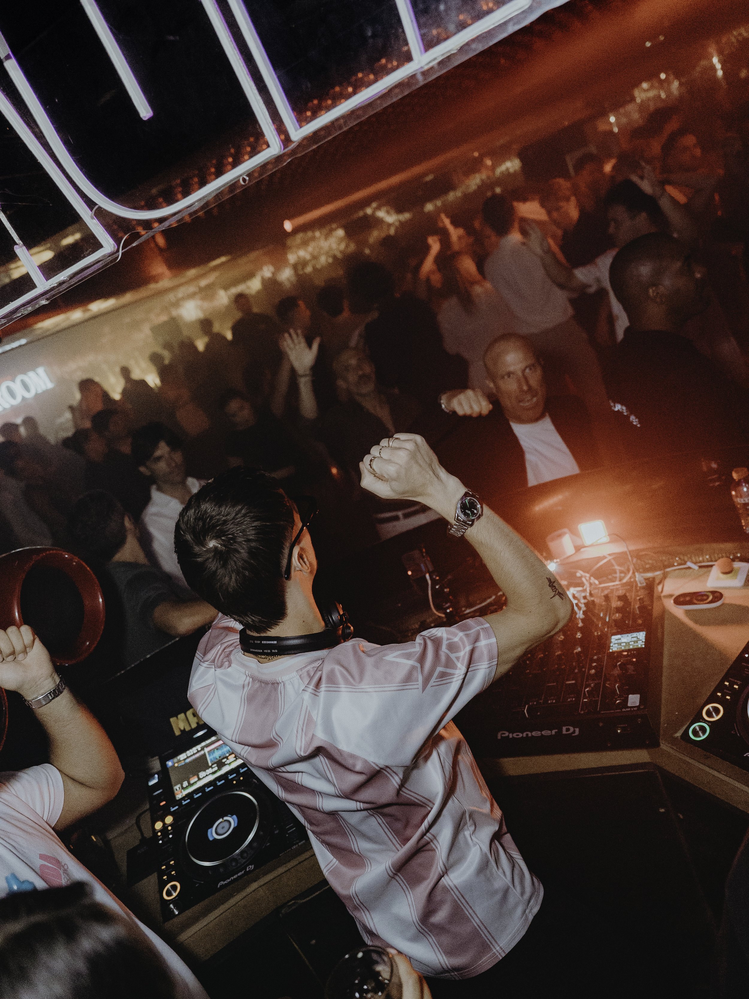 DJ performan dans une boîte de nuit bondée, les gens dansent et socialisent sous un éclairage rouge.