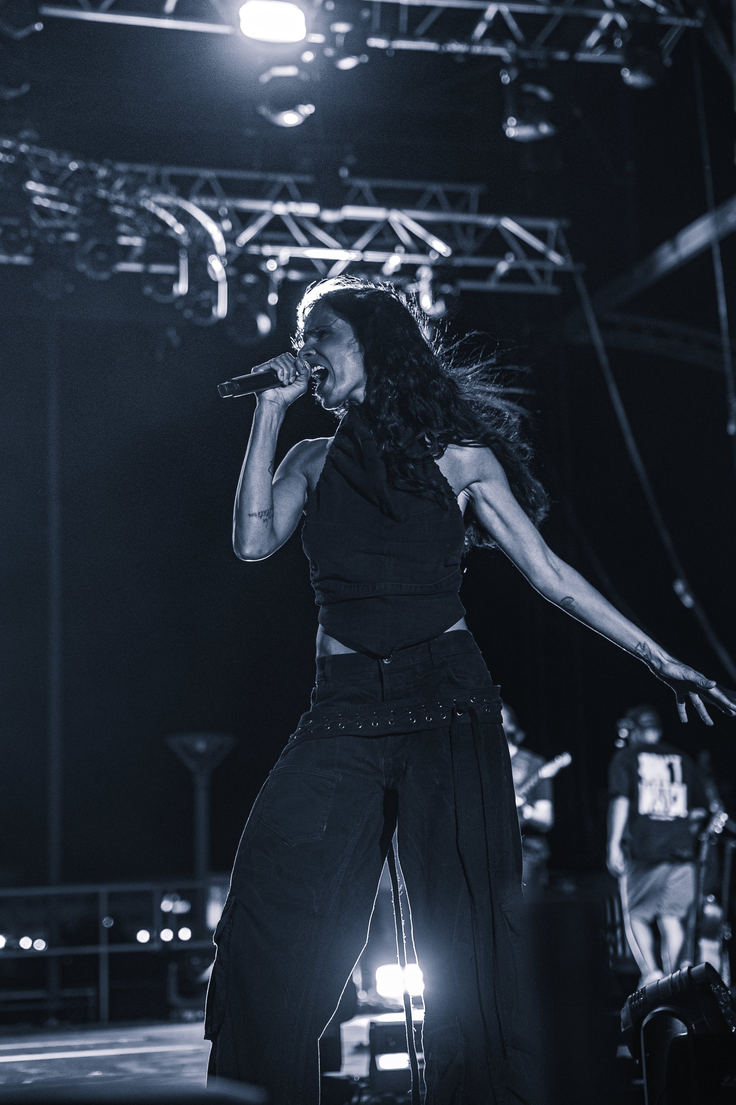 Une chanteuse avec des cheveux longs et bouclés chante dans un microphone lors d'un concert, version en noir et blanc avec un fond de scène avec des dispositifs d'éclairage.