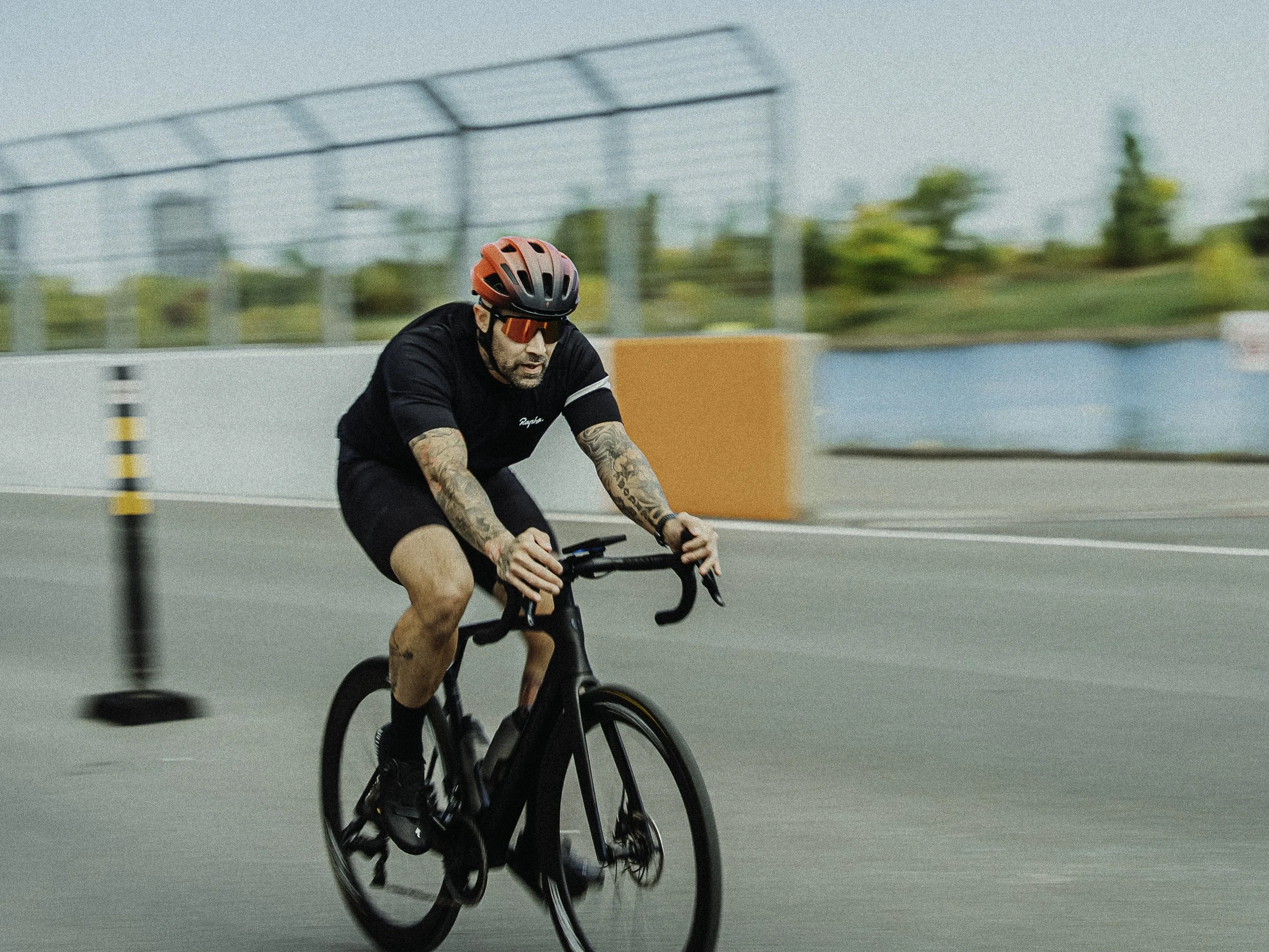Un homme fait du vélo sur une piste urbaine, portant un casque rouge et des lunettes de soleil, avec des tatouages sur les bras.