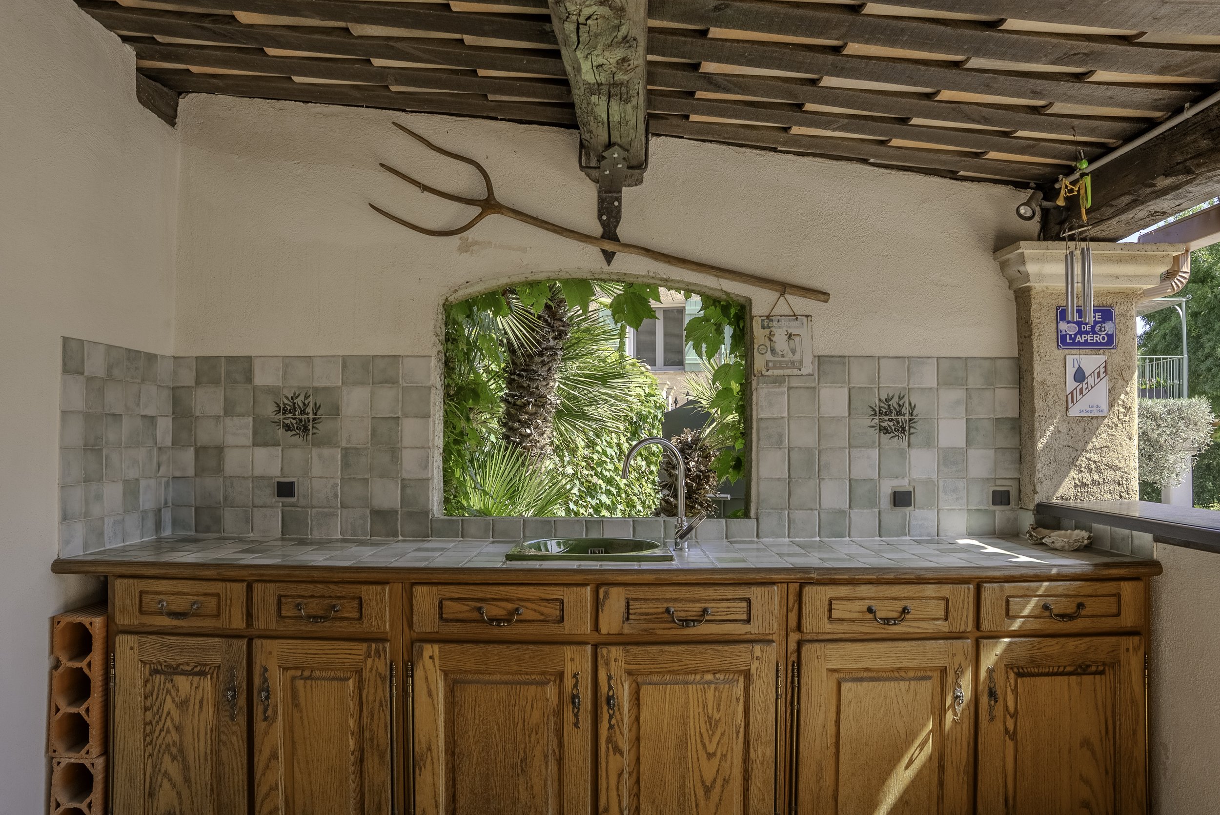 Coin de cuisine avec un comptoir en bois, un évier, un plafond en bois avec une faucille suspendue, et une ouverture donnant sur un jardin avec des plantes et un palmier.
