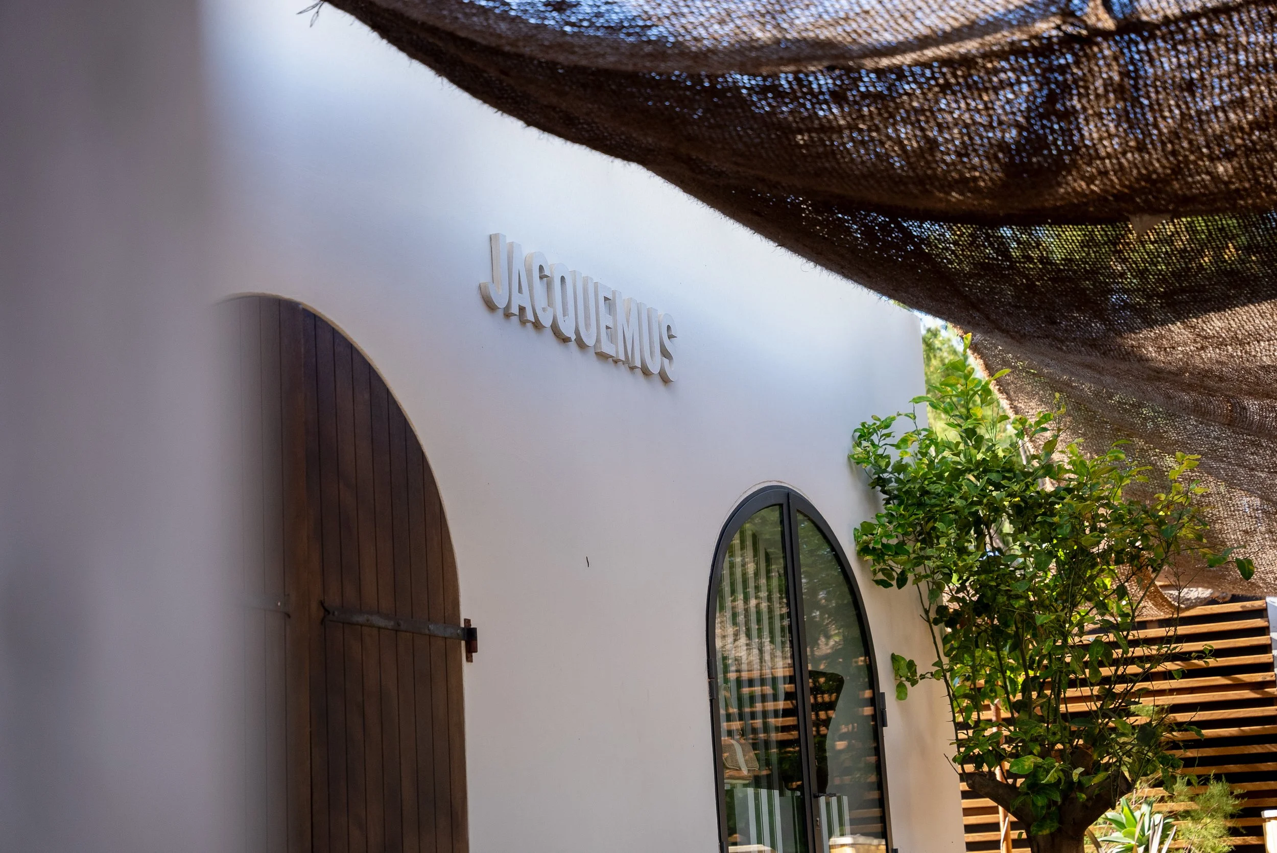 Façade d'un bâtiment blanc avec une fenêtre en arc et une porte en bois, le nom "JACQUEMUS" en lettres majuscules sur le mur, une petite arbre vert à côté, une couverture en tissu noir en haut.