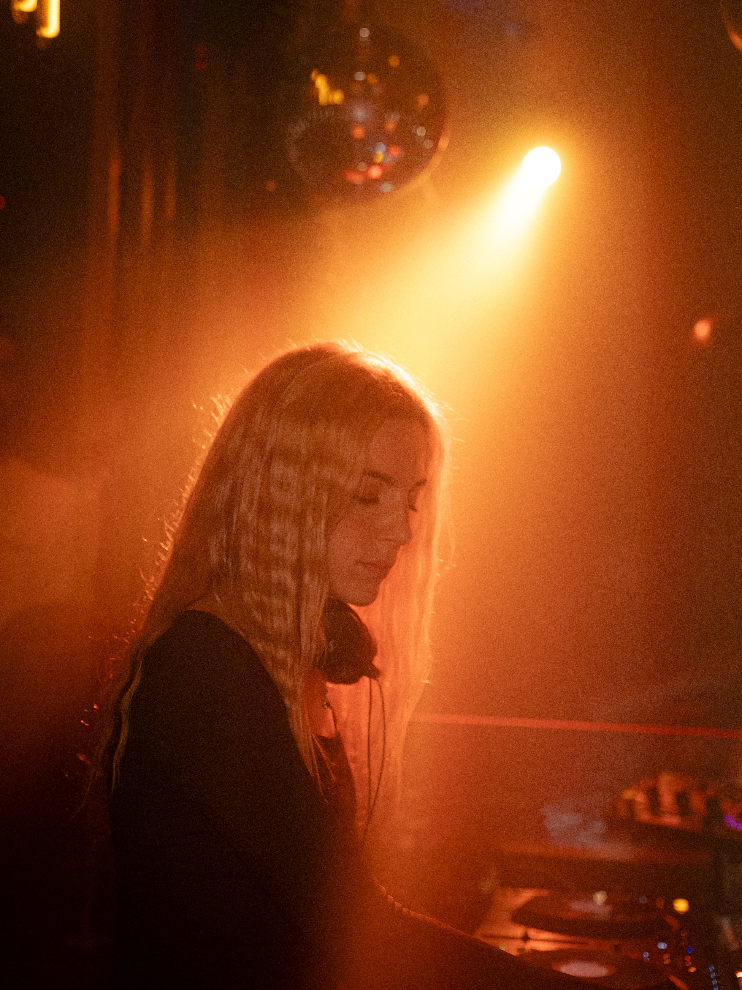 Jeune femme DJ avec des cheveux longs blonds, portant des écouteurs, en train de mixer dans une discothèque, sous un éclairage orange et jaune, avec une boule disco en haut.