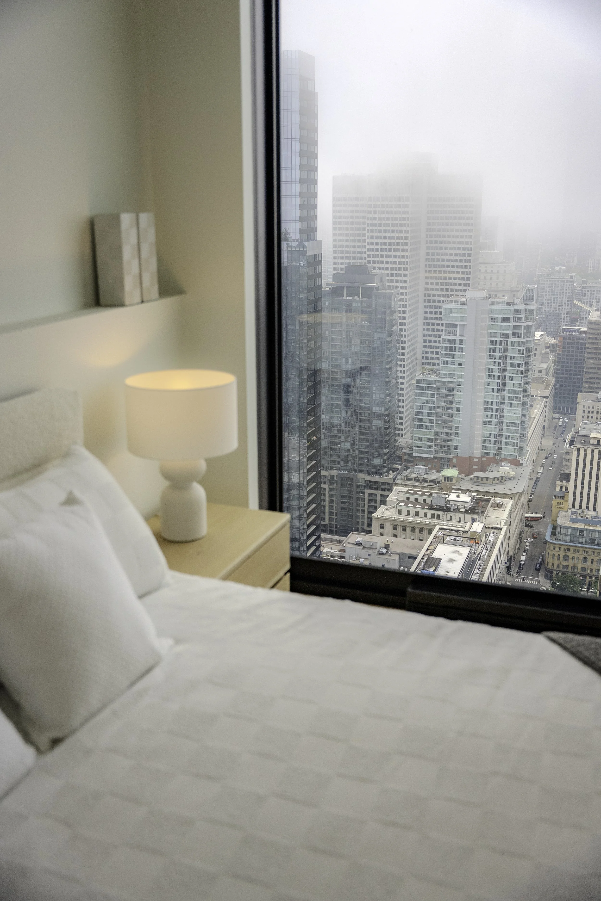Chambre avec lit blanc, lampe de chevet, vue sur une ville avec de nombreux gratte-ciel sous la brume