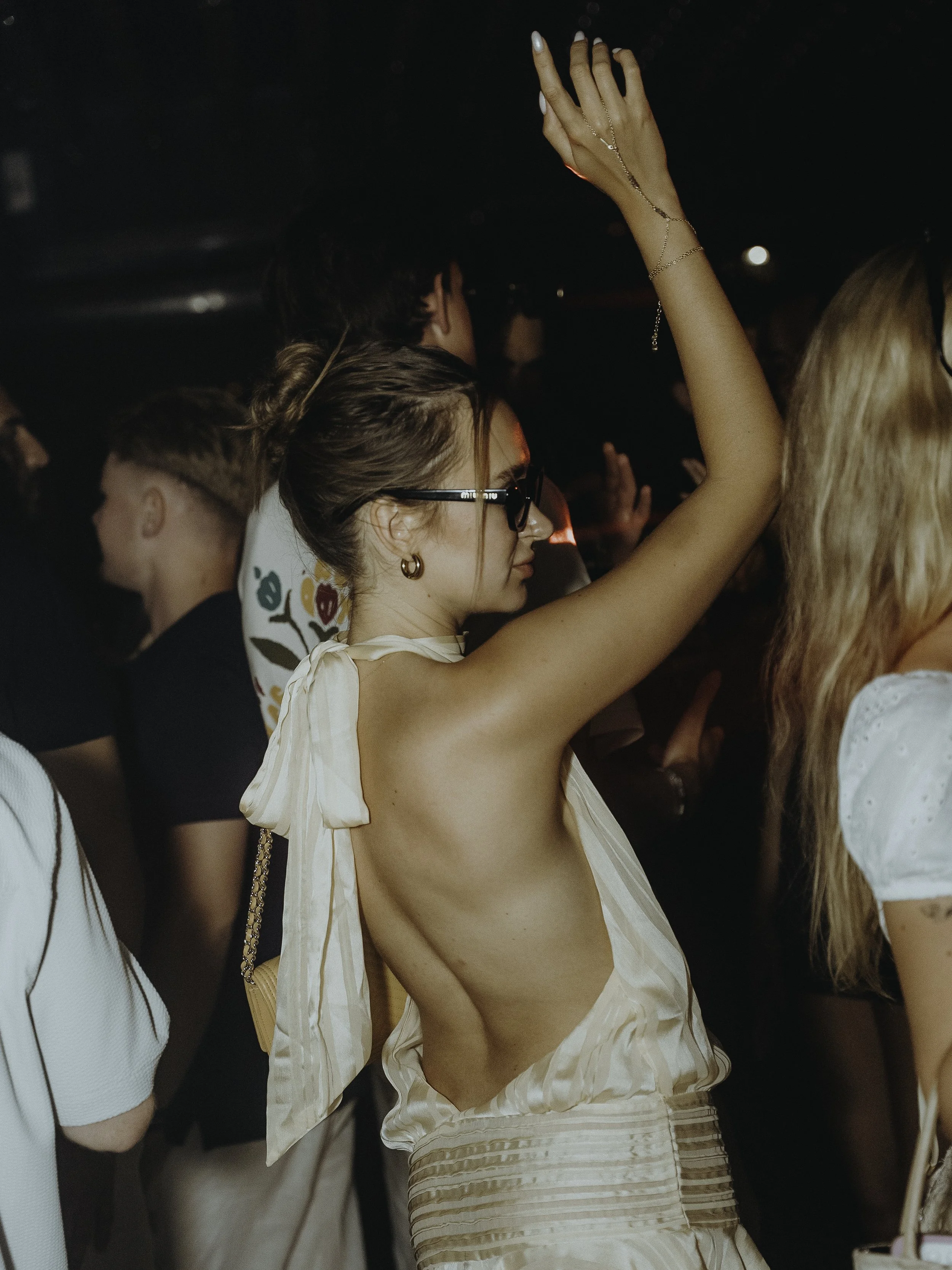 Femme dansant dans une fête ou un club, portant une robe beige ouverte dans le dos, portant des lunettes noires et des bijoux.