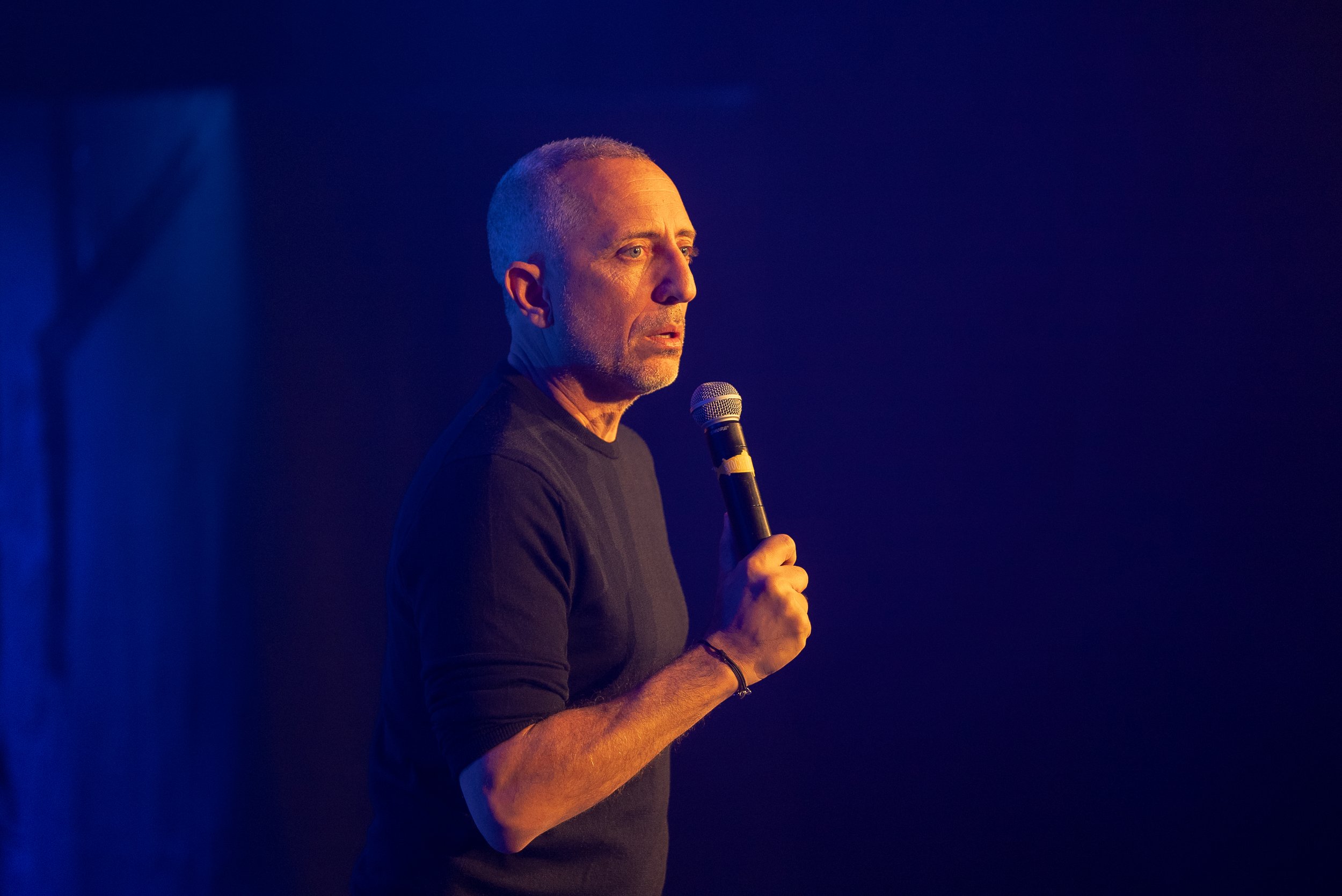 Un homme gris tenant un microphone lors d'une performance ou d'un discours, dans un éclairage chaud et sombre.