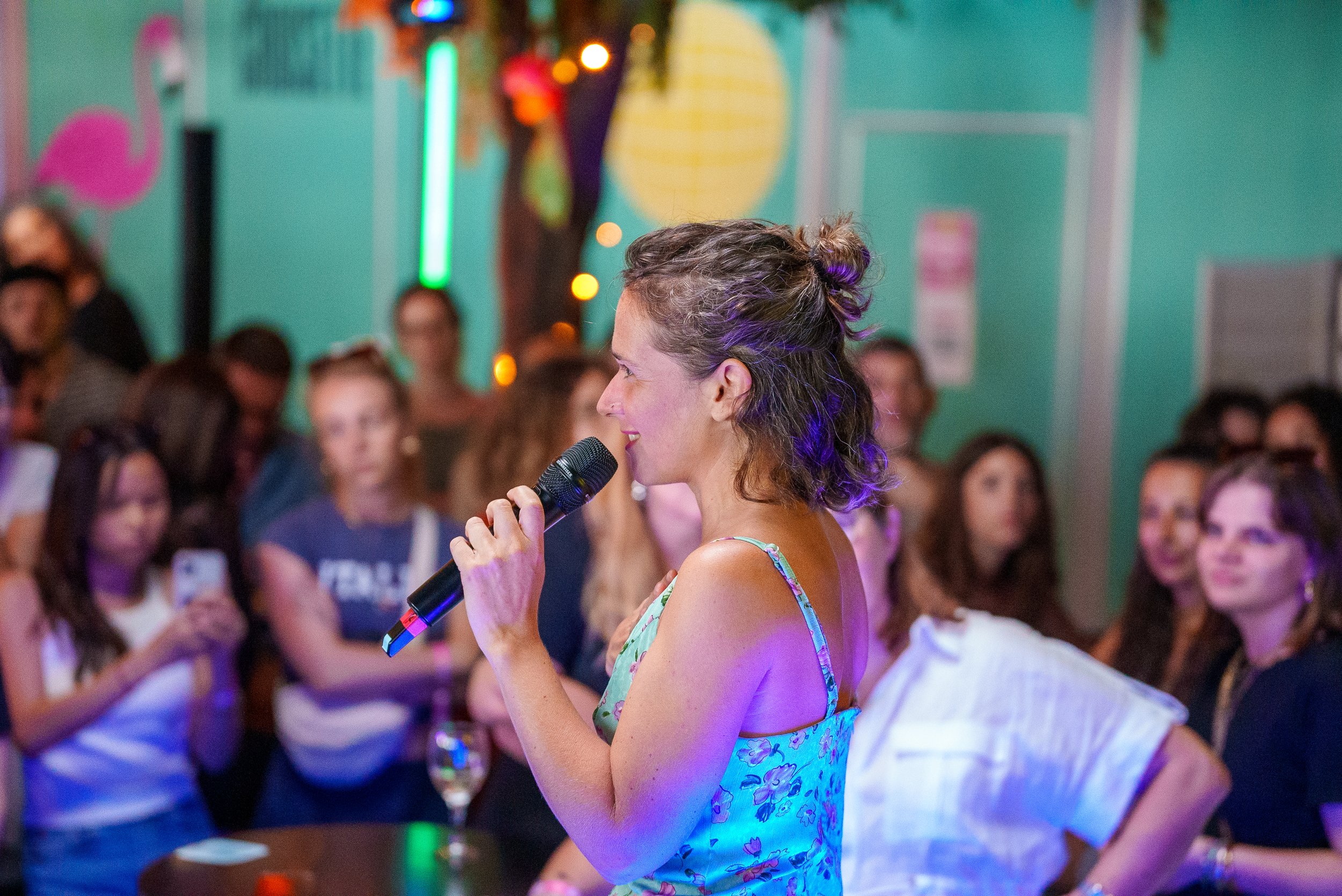 Une femme parle dans un micro lors d'une soirée dans un lieu décoré de néons, entourée d'un public debout qui l'écoute attentivement.