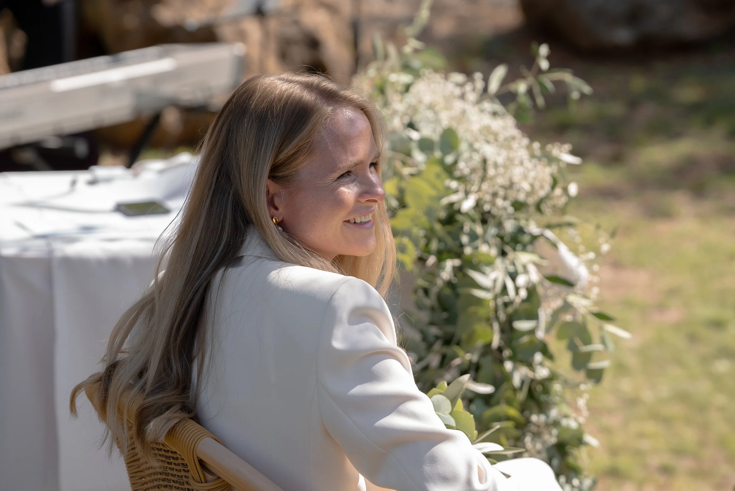 Une femme blonde souriante en tenue blanche, assise à l'extérieur près de verdure et de fleurs, regardant vers la droite.