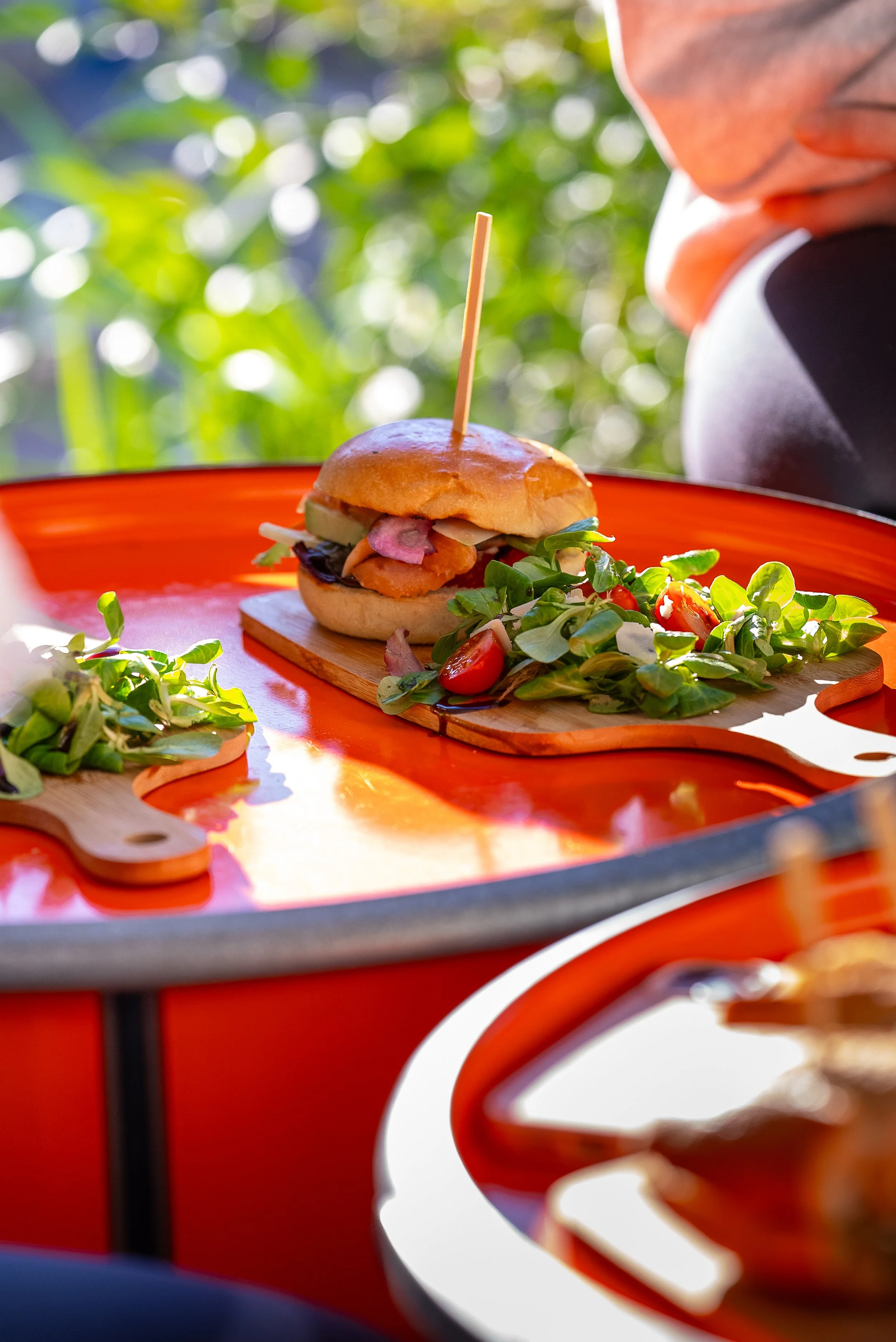 Un sandwich avec légumes frais et une salade à côté, présenté sur une planche en bois, sur une grande plateforme orange avec un fond flou de feuillage vert.
