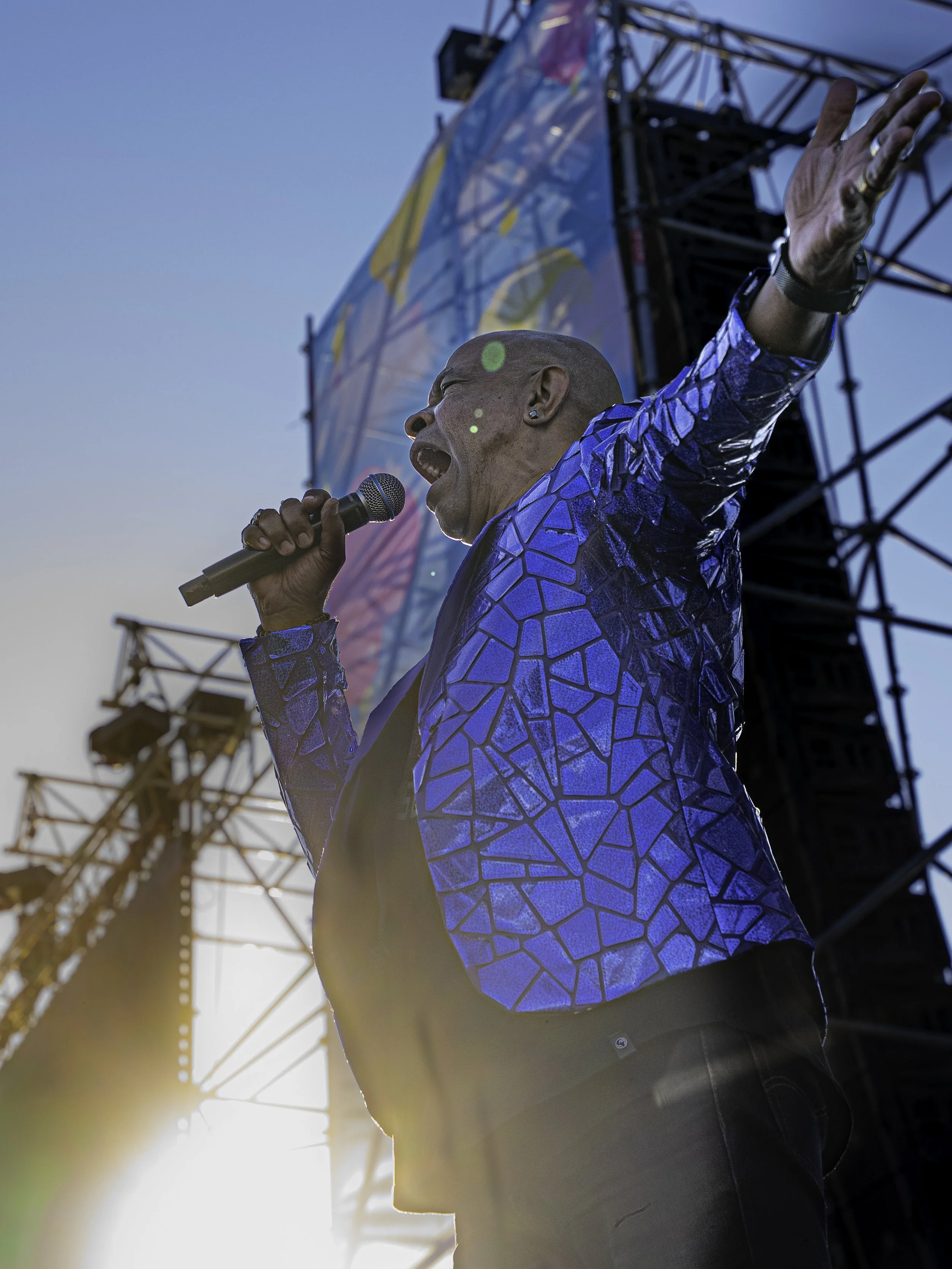 Un homme chantant avec un micro lors d'un concert en plein air, portant une veste bleue avec un motif géométrique, avec la scène et le ciel en arrière-plan.