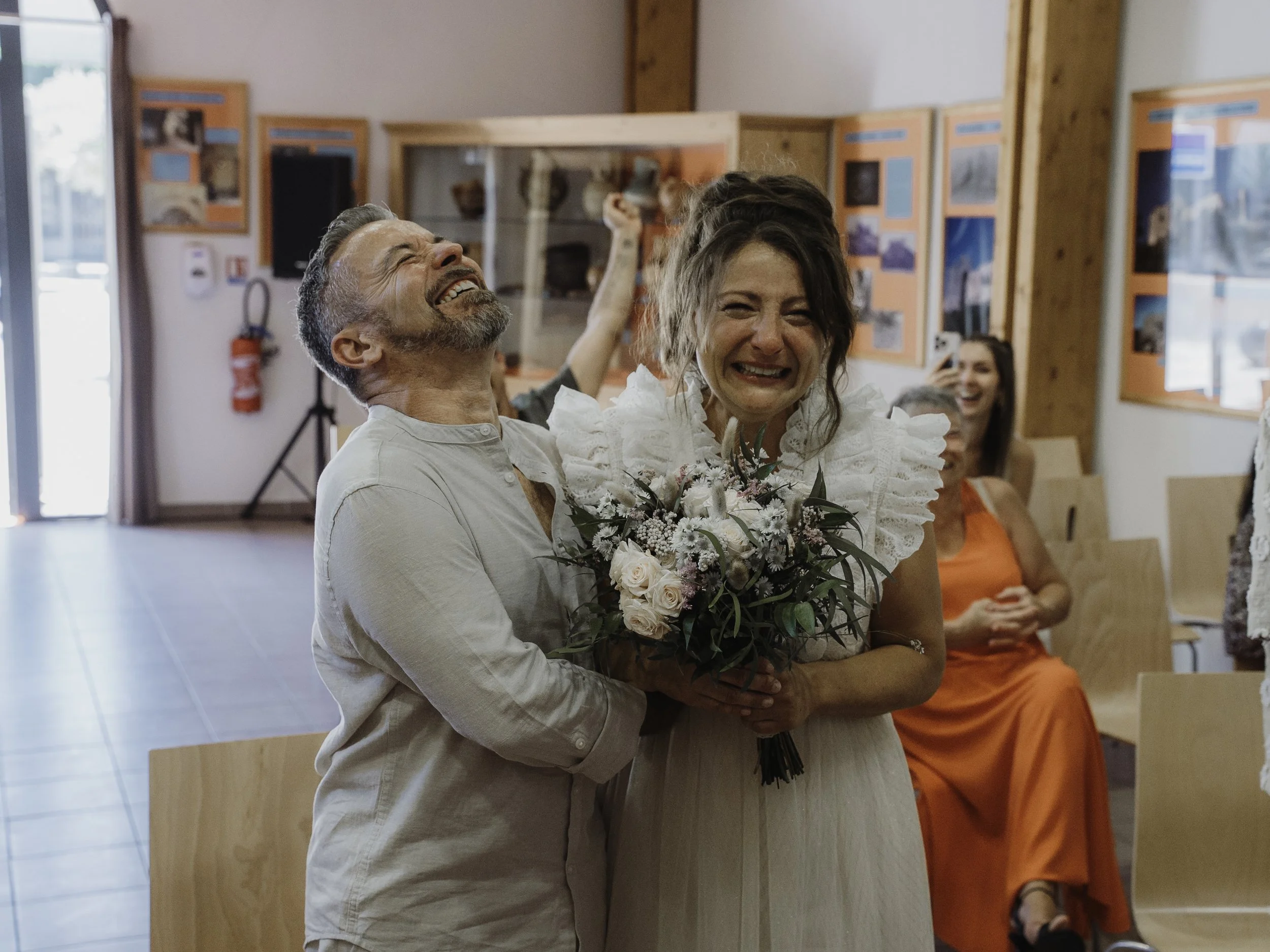 Une femme en robe blanche tenant un bouquet de fleurs, visiblement émue, est entourée de personnes qui semblent célébrer un mariage ou un moment festif, dans une salle avec des affiches en fond.