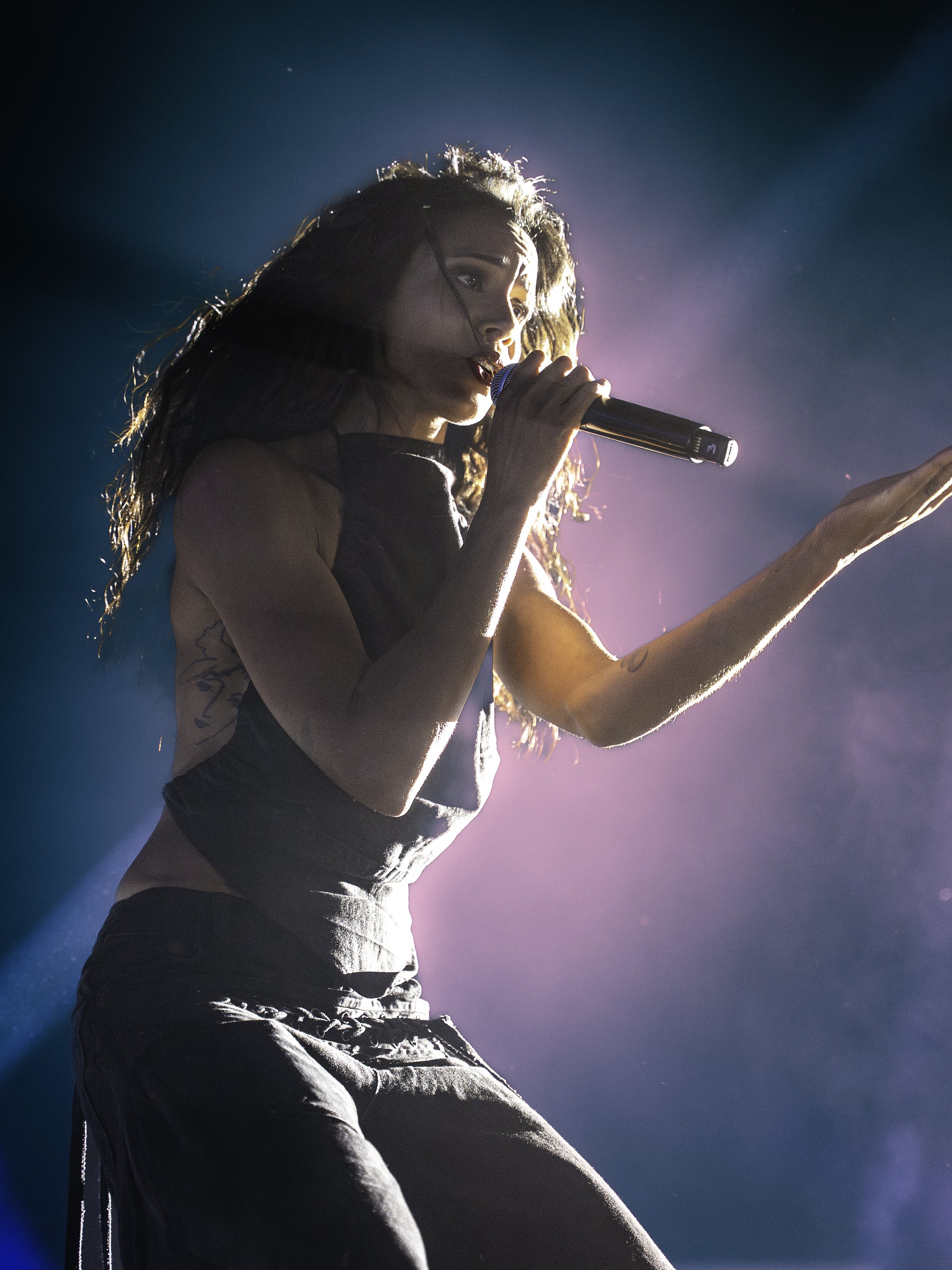 Une femme chante avec un microphone sur scène, éclairée par des lumières colorées, portant un haut sans manches en noir et un pantalon en tissu
