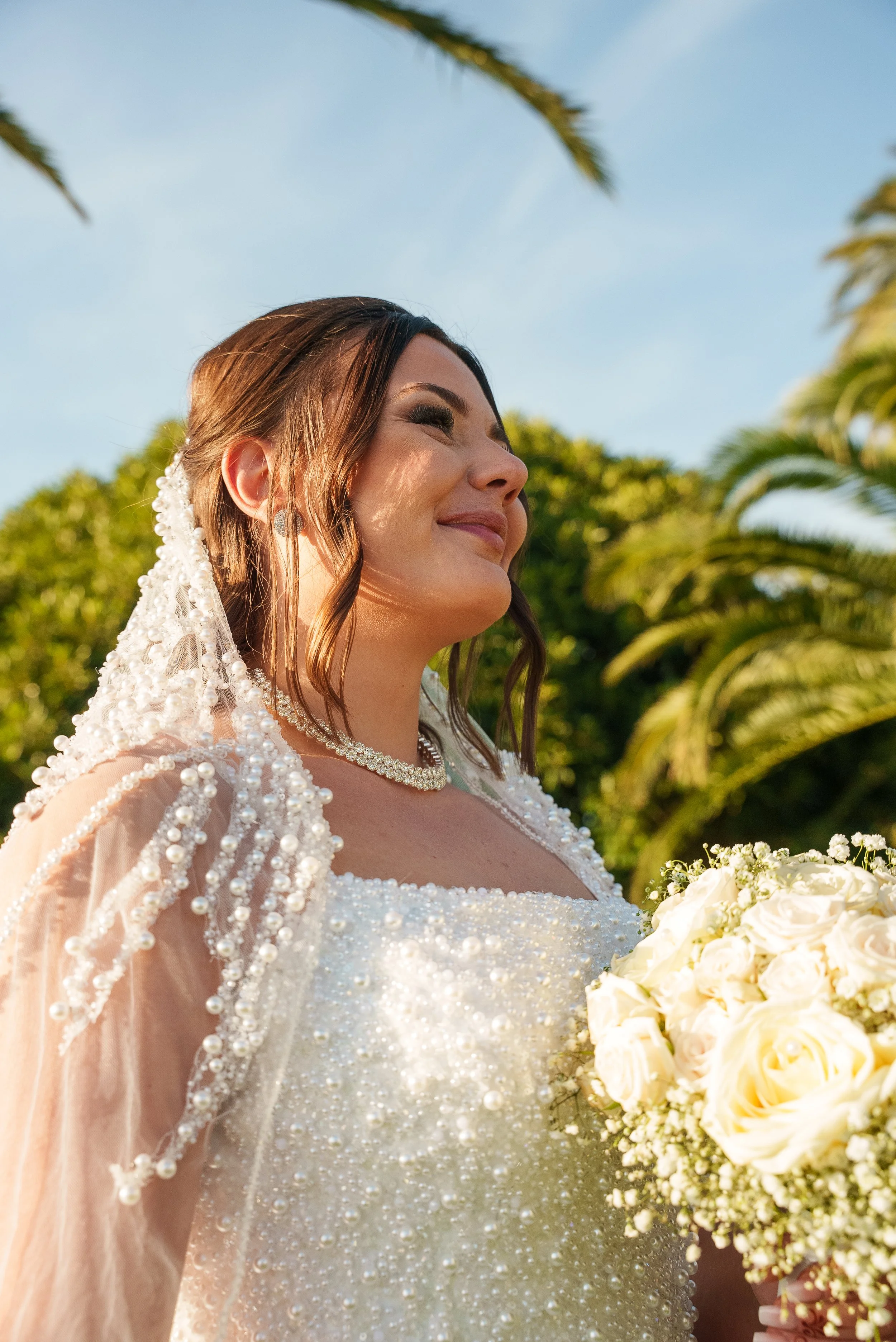 Une femme en robe de mariée sourit, tenant un bouquet de roses blanches, dans un environnement en plein air avec des sapins et un ciel bleu.