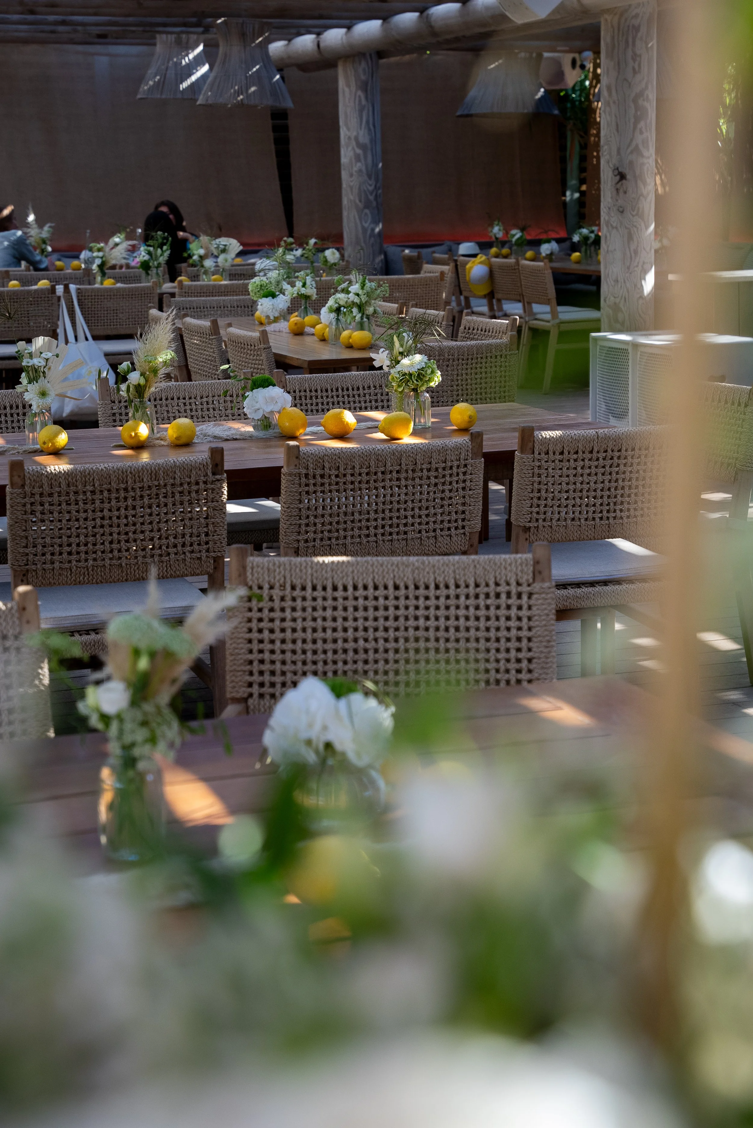 Tables décorées avec des fleurs et des citrons jaunes, assises en extérieur sous un toit en bois, avec des chaises en rotin.