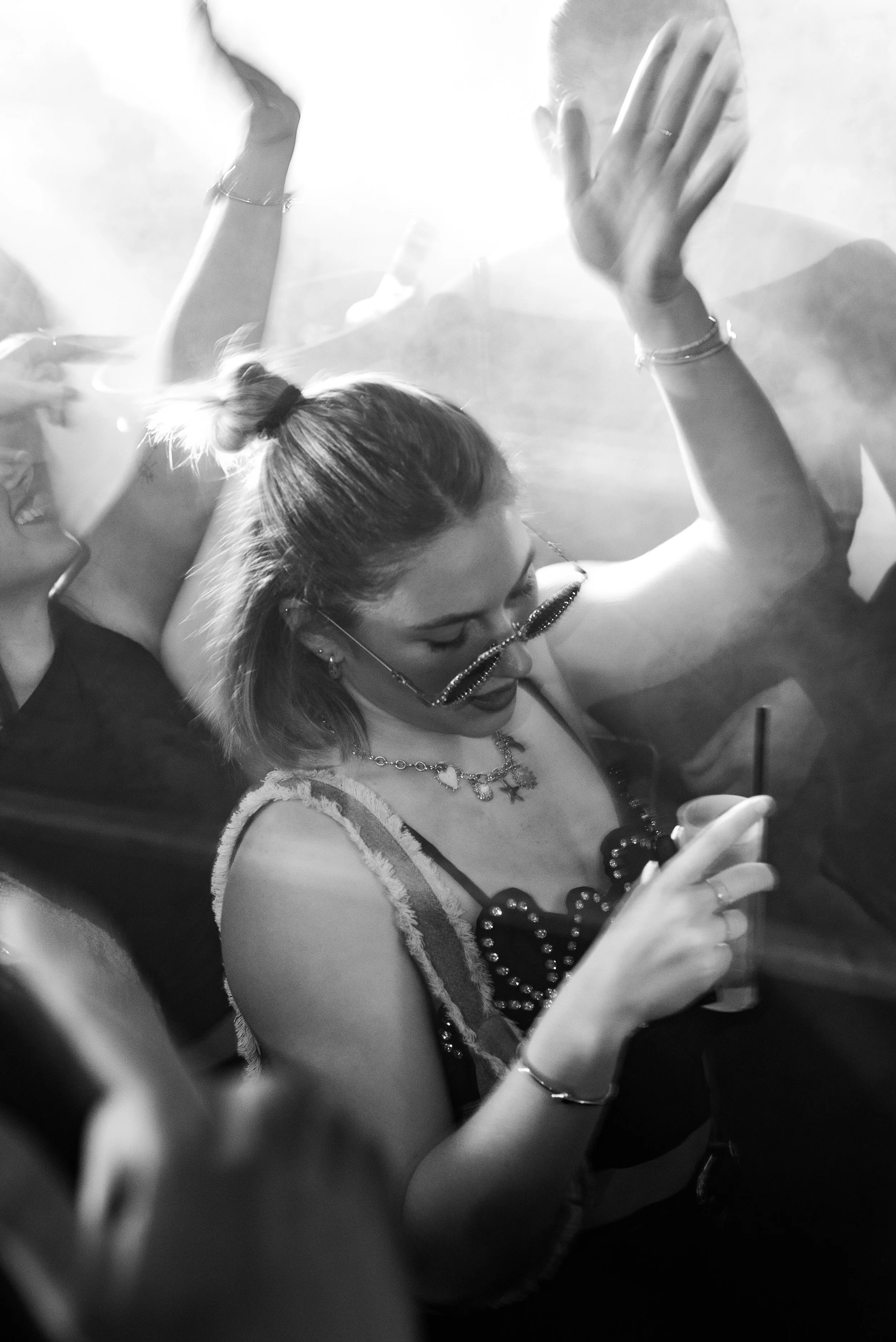 Femme jeune avec lunettes de soleil et bijoux, regardant son téléphone dans une ambiance de fête ou discothèque, en noir et blanc.